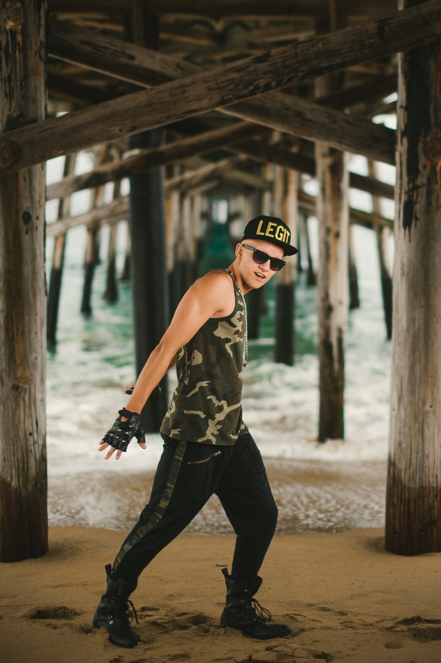 Sammy C Side profile of man standing under a pier wearing a camo shirt, black pants and military boots, black cap with LEGIT written on it in gold and sunglasses posing for the camera