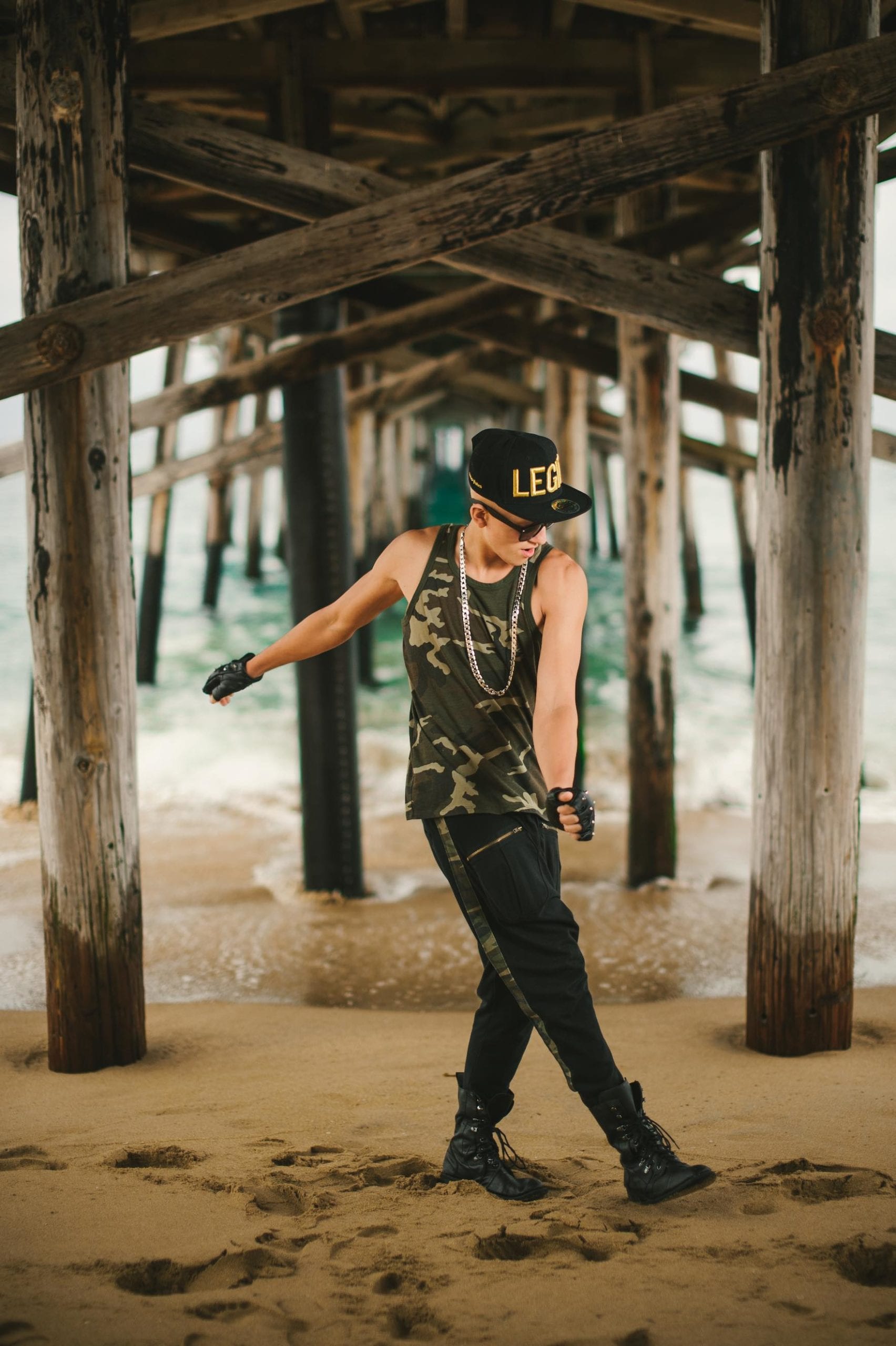 Sammy C Side profile of man standing under a dock wearing a camo shirt, black pants and military boots, black cap with LEGIT written on it in gold and sunglasses posing for the camera looking off screen