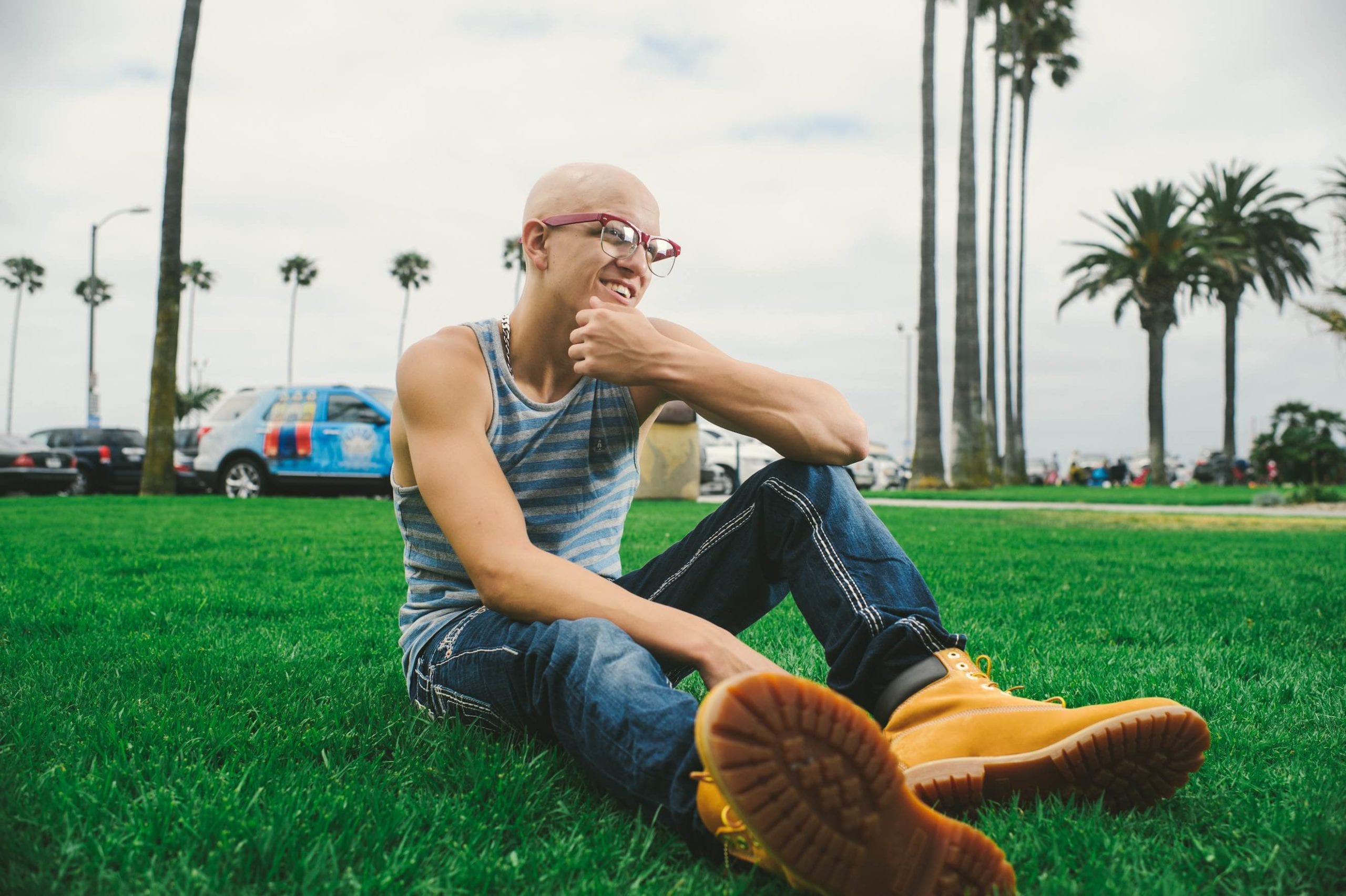 Sammy C Bald man wearing blue and white striped tank top, blue jeans, red glasses and beige work shoes sitting in the grass with palm trees and cars in the background