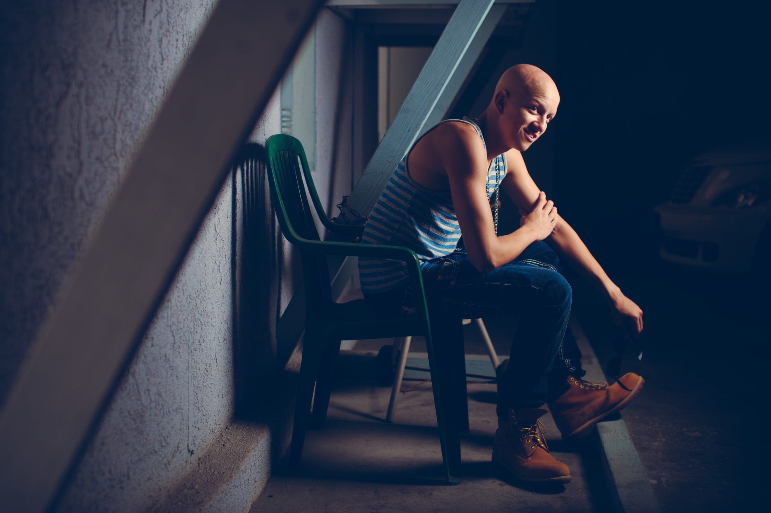 Bald young man sitting on a green plastic chair facing the camera sideways. He is wearing a blue and white striped tank top with metal necklace along with leather workman's boots.