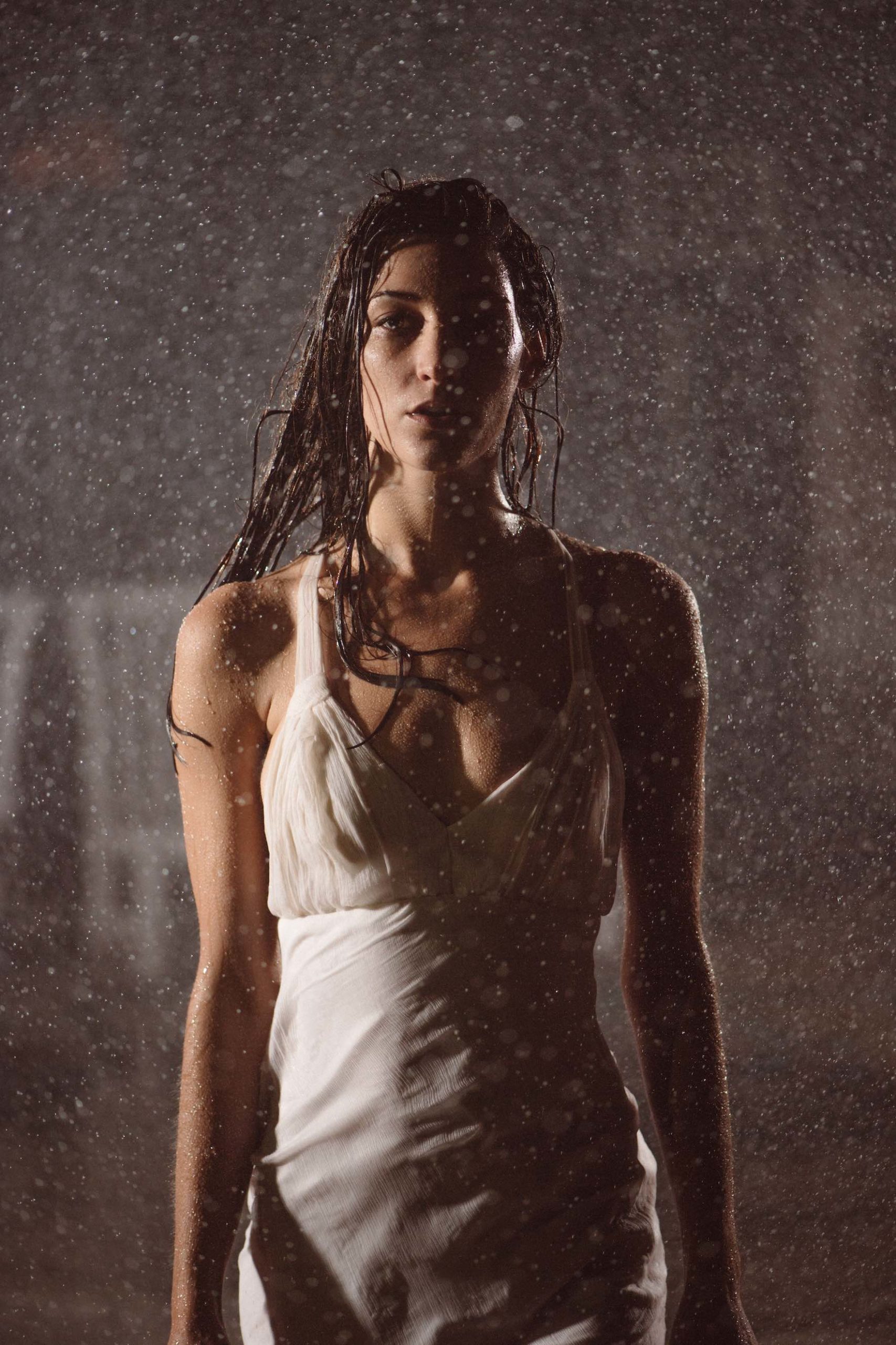 Woman with long hair wearing a white dress posing for camera under a shower of water