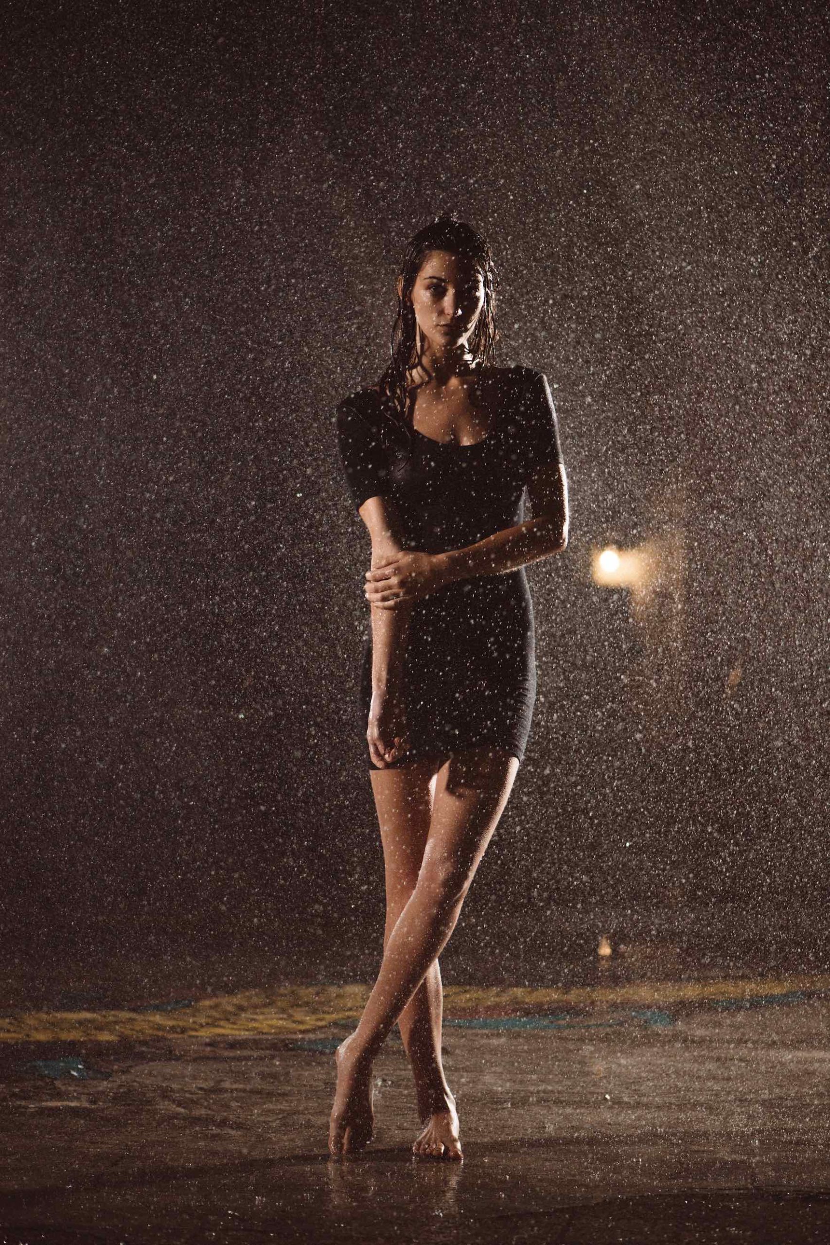 Woman with long hair posing for camera barefoot wearing a black dress under a shower of water