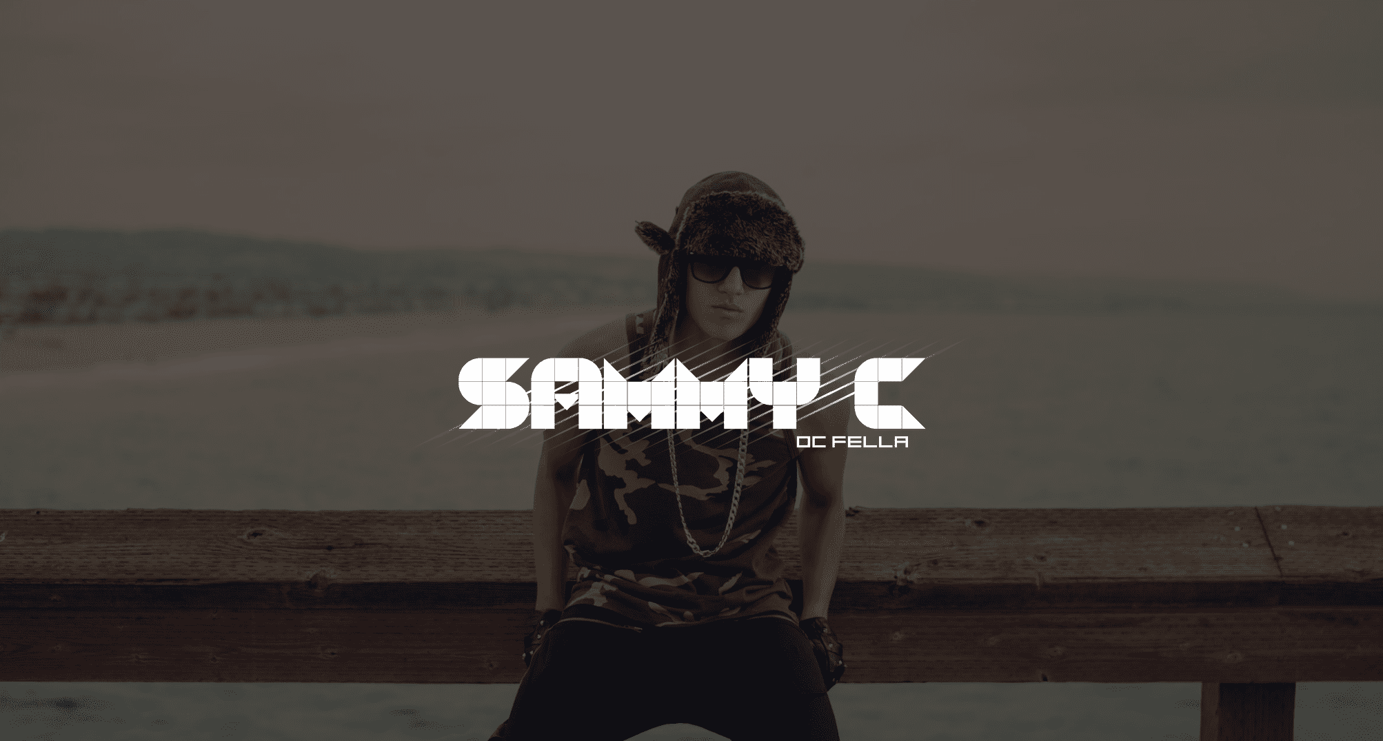 White Sammy C DC Fella logo with background of young man wearing black pants and camouflage tank top with black shoes and shades posing for camera with black leather gloved hands at his sides. He is sitting on a wooden railing by the ocean.