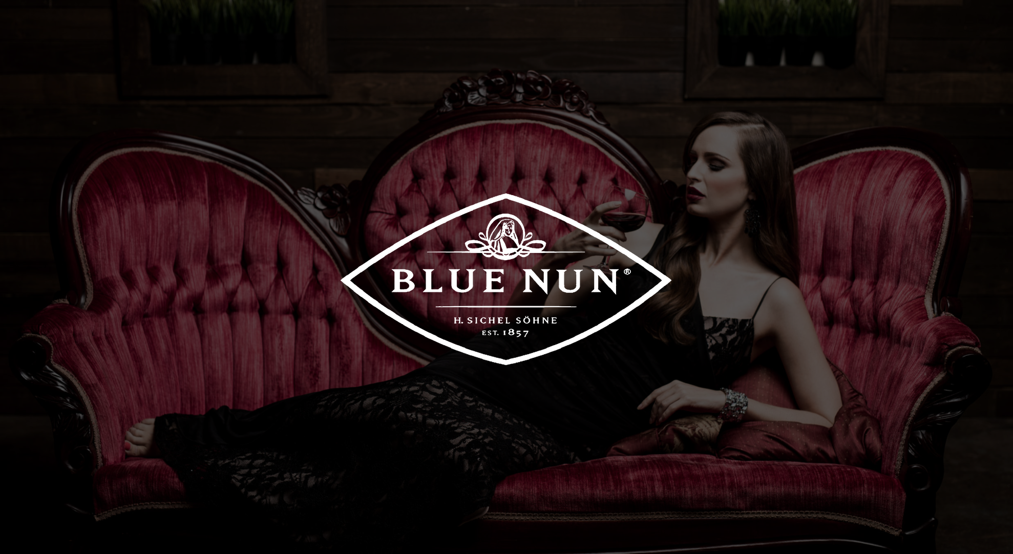 White Blue Nun logo with dimmed background of woman laying on a vintage red couch holding a glass of wine posing for the camera