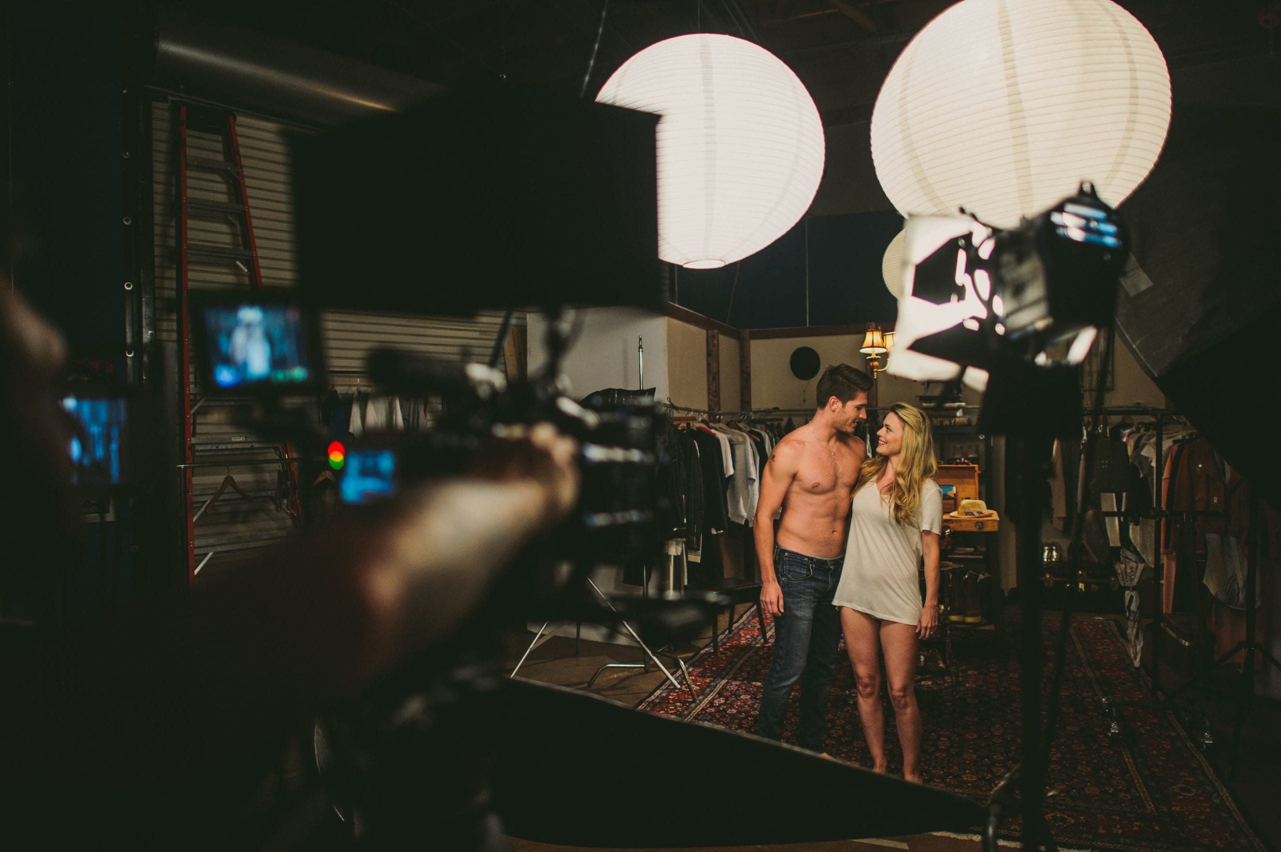 Behind the scenes look at a shirtless man with a woman wearing a long gray t shirt being filmed by videographer in a studio