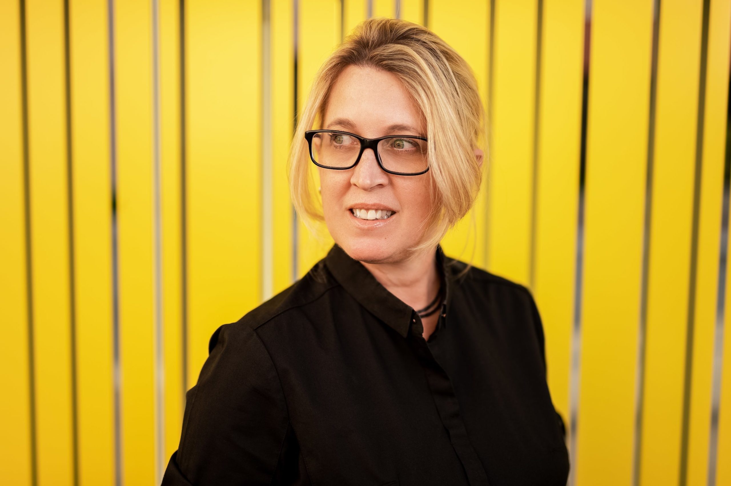 Headshot of woman with short blond hair and glasses wearing a black dress shirt posing for camera in front of yellow wall looking off to the side