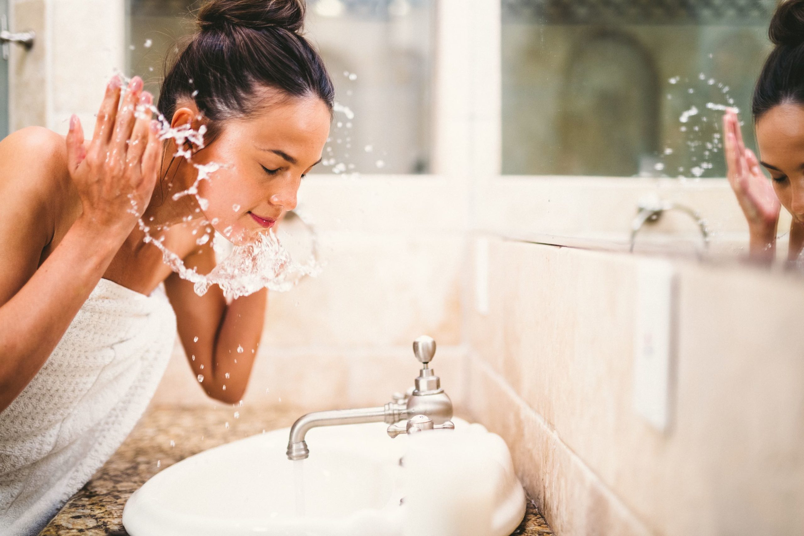 Woman splashing water on her face by a sink with a mirror showing her reflection