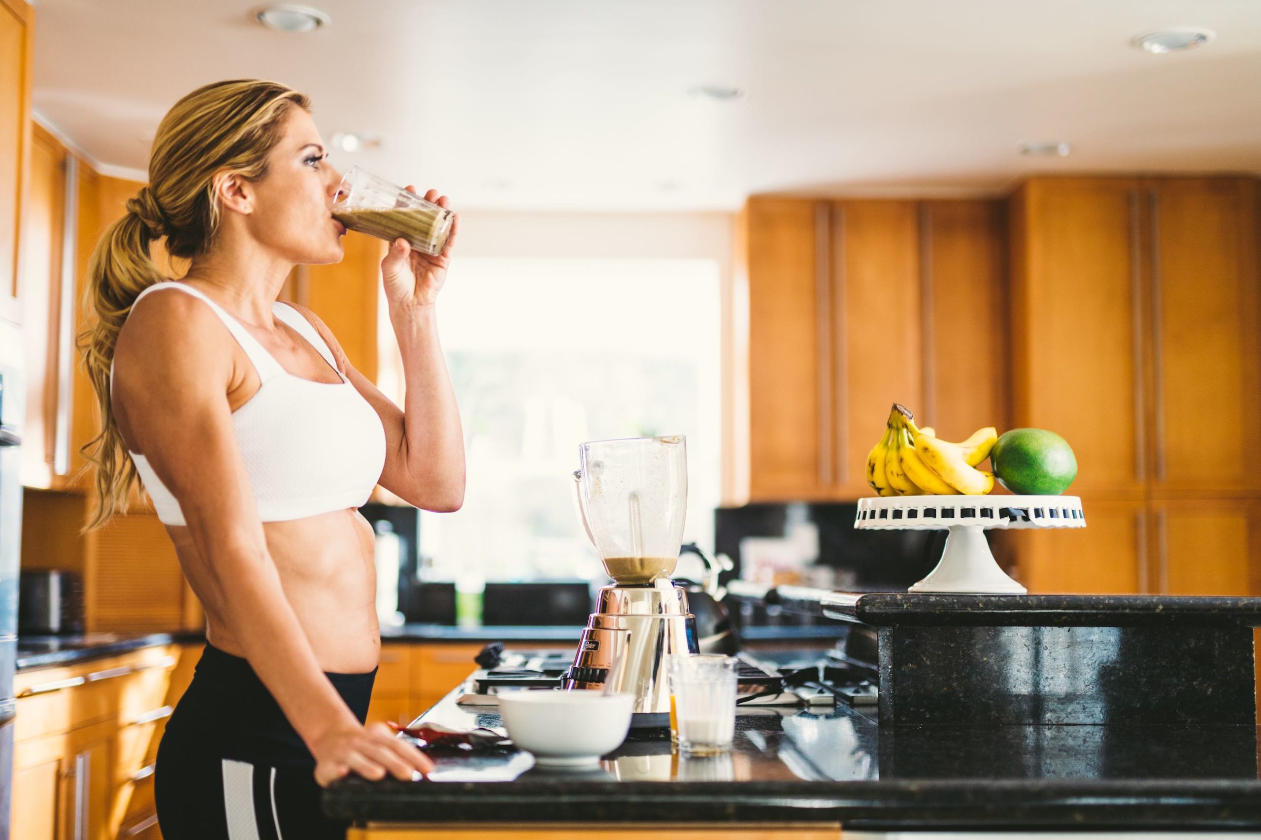 Side profile of woman wearing white sports bra and black pants drinking a smoothie in a kitchen by a mixer on a counter