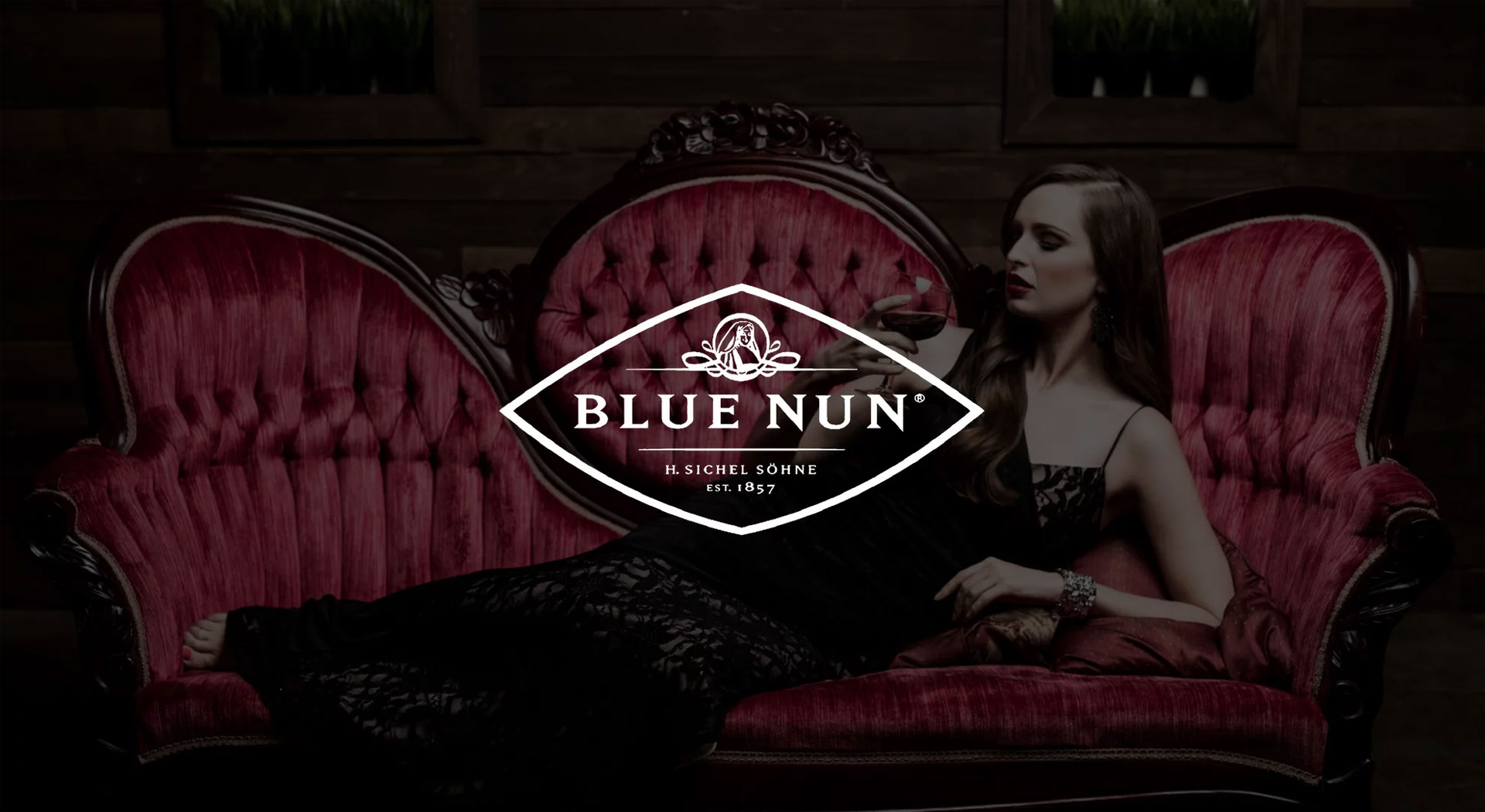 IU C&I Studios Page White Blue Nun logo with dimmed background of woman laying on a vintage red couch holding a glass of wine posing for the camera