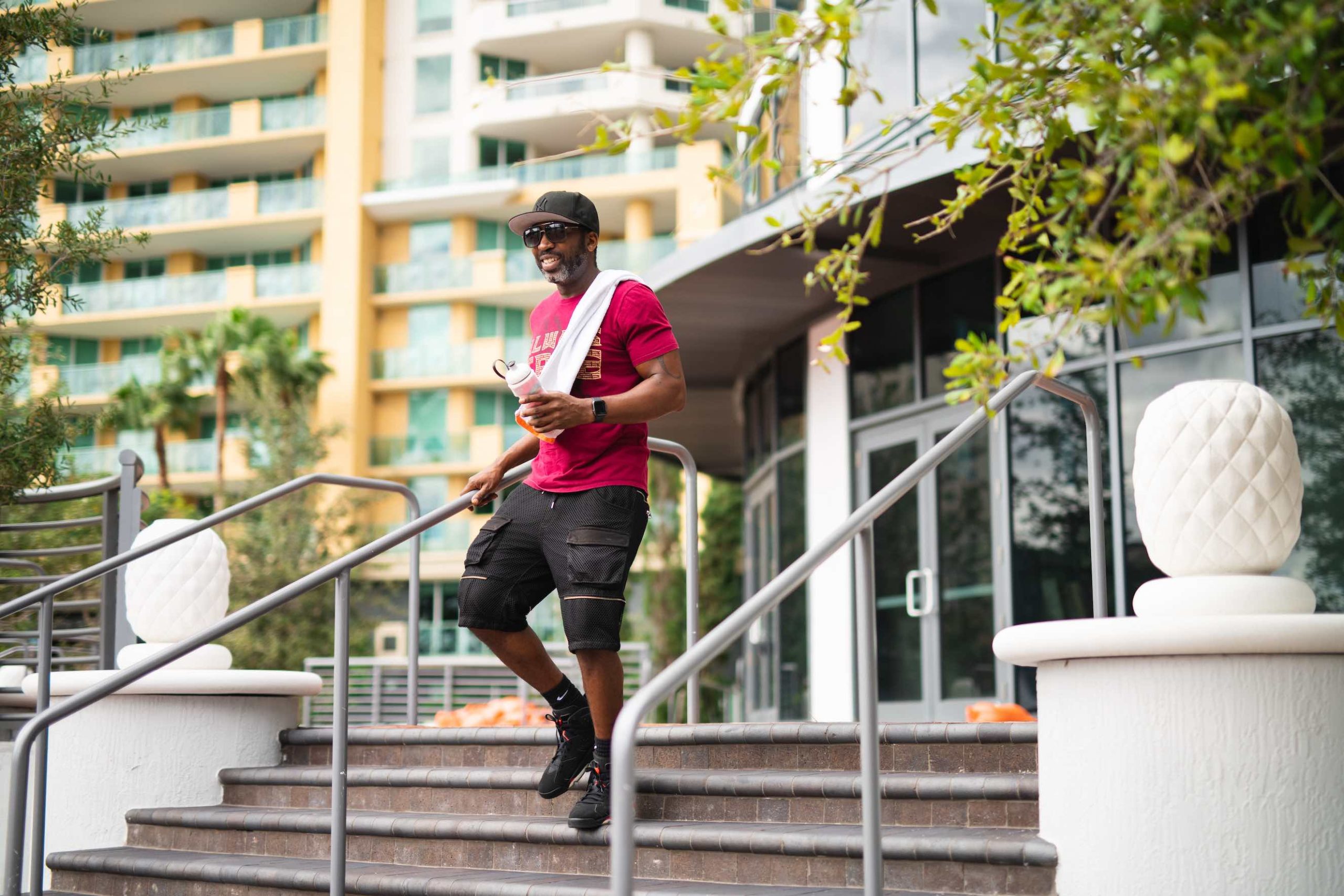 Freebird Real Estate East Fort lauderdale African American man wearing black cap, shades, salt and pepper goatee, red shirt and black shorts walking down steps holding a water bottle