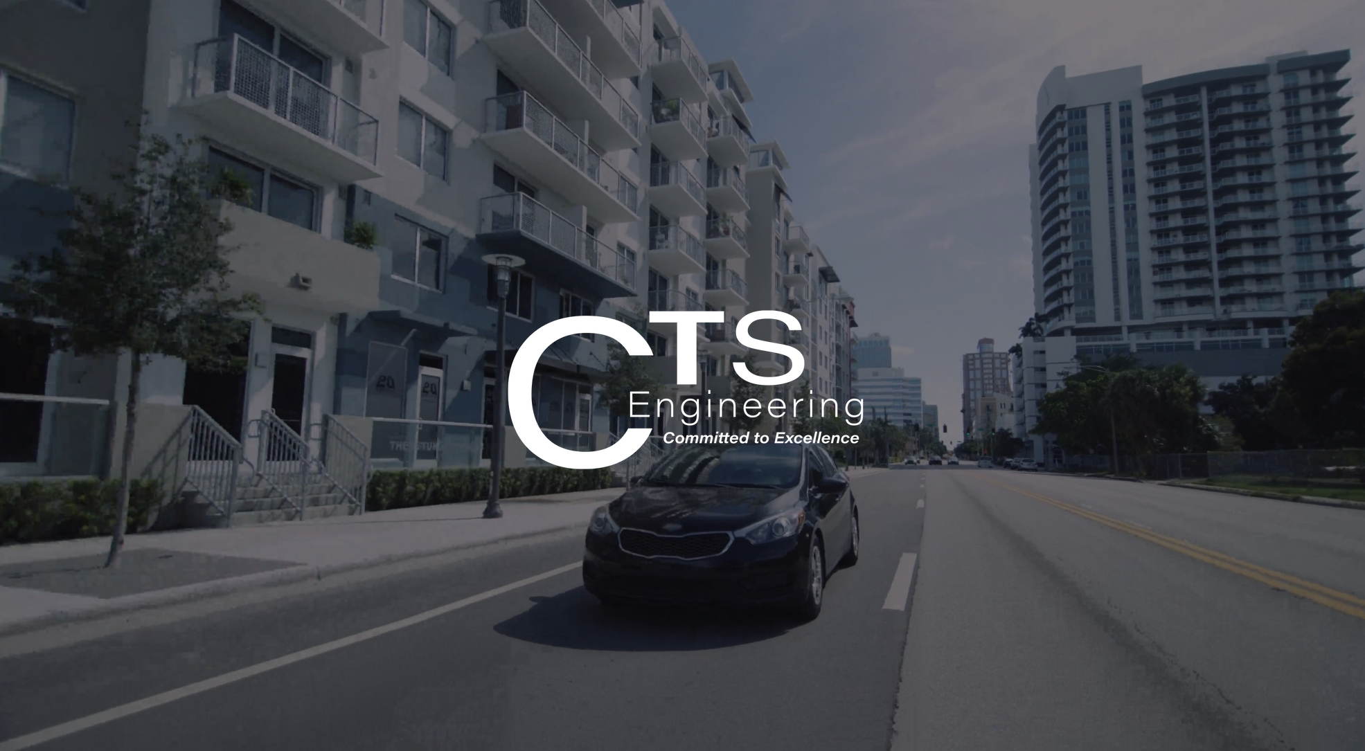 White CTS Engineering Committed to Excellence logo with dimmed background view from front of a black car driving down a road in a city