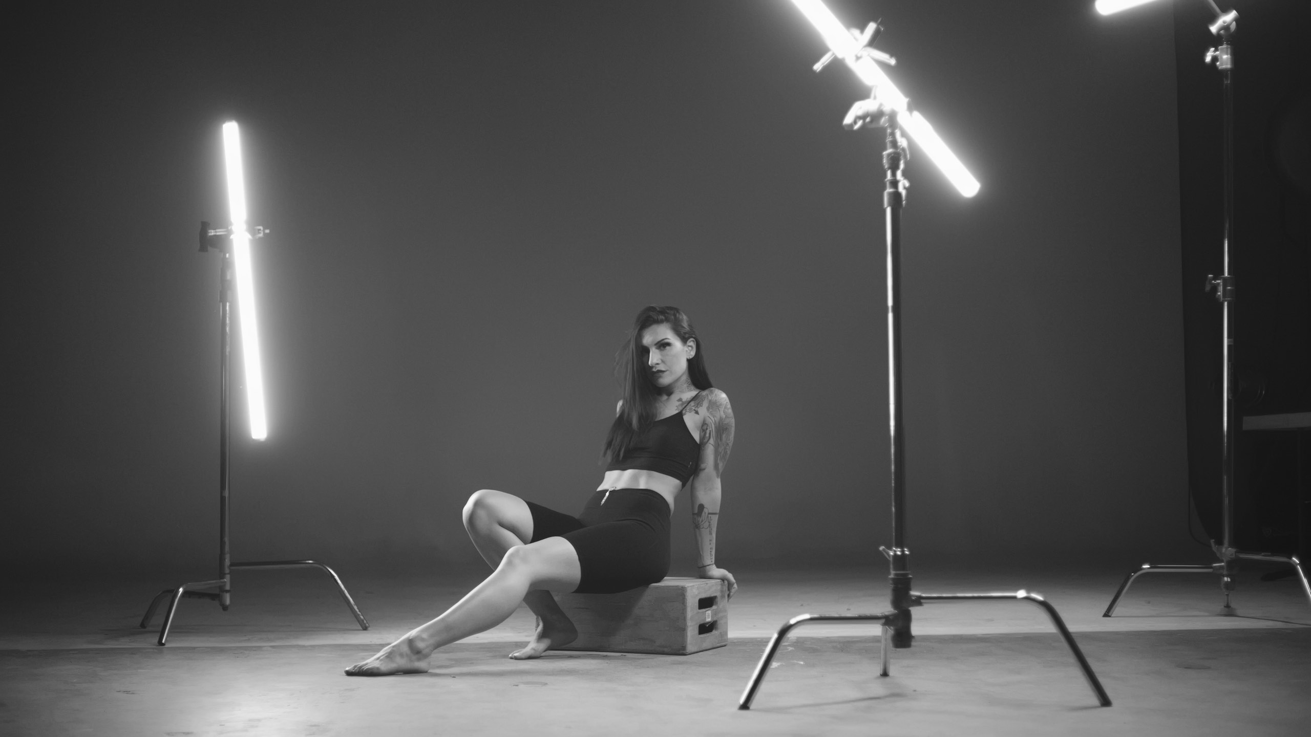 IU Puma Black and white side profile of tattooed woman in in black top and shorts under lights sitting on a wooden box