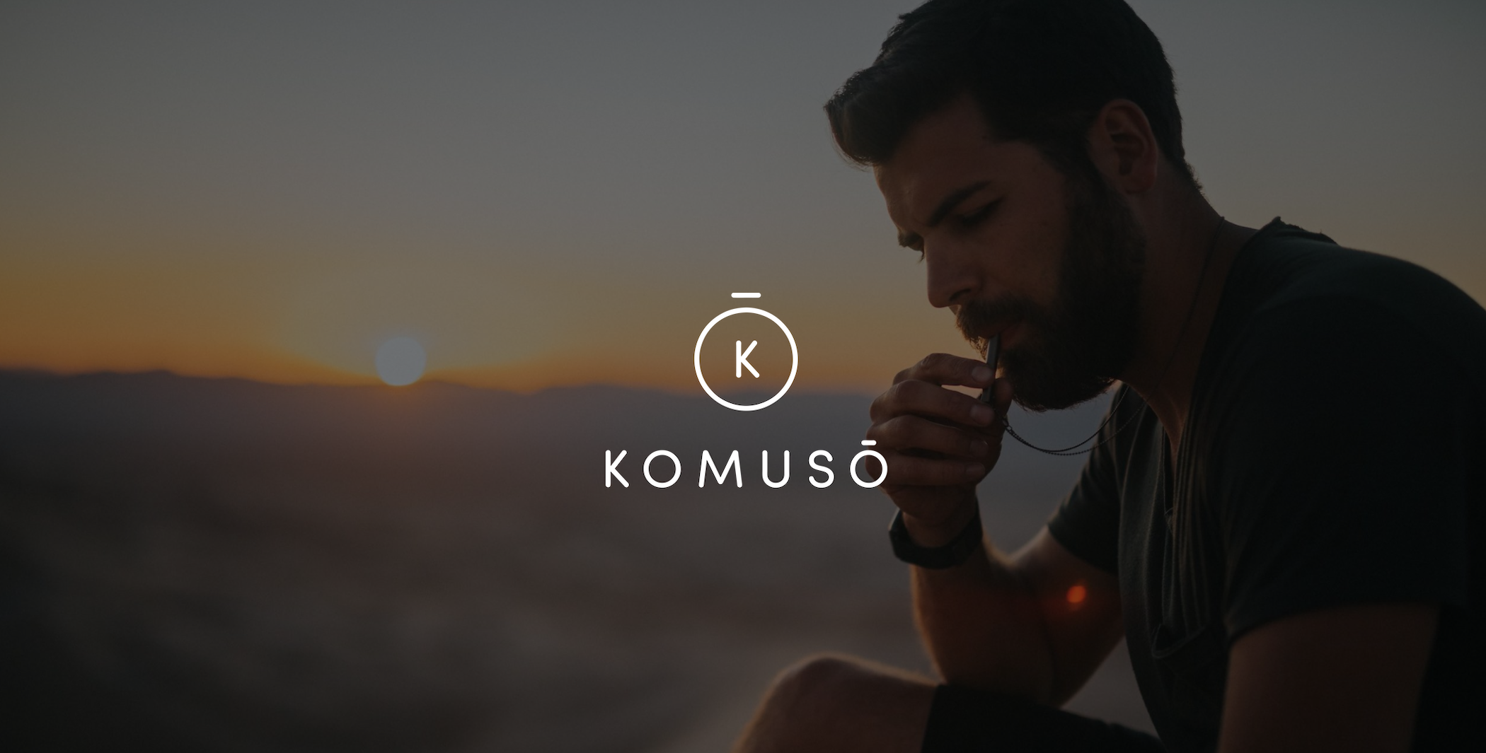 IU C&I Studios Page Branding services for Komuso Design White Komuso logo with dimmed background of a bearded man blowing a whistle with a view of the sun setting in the valley behind him