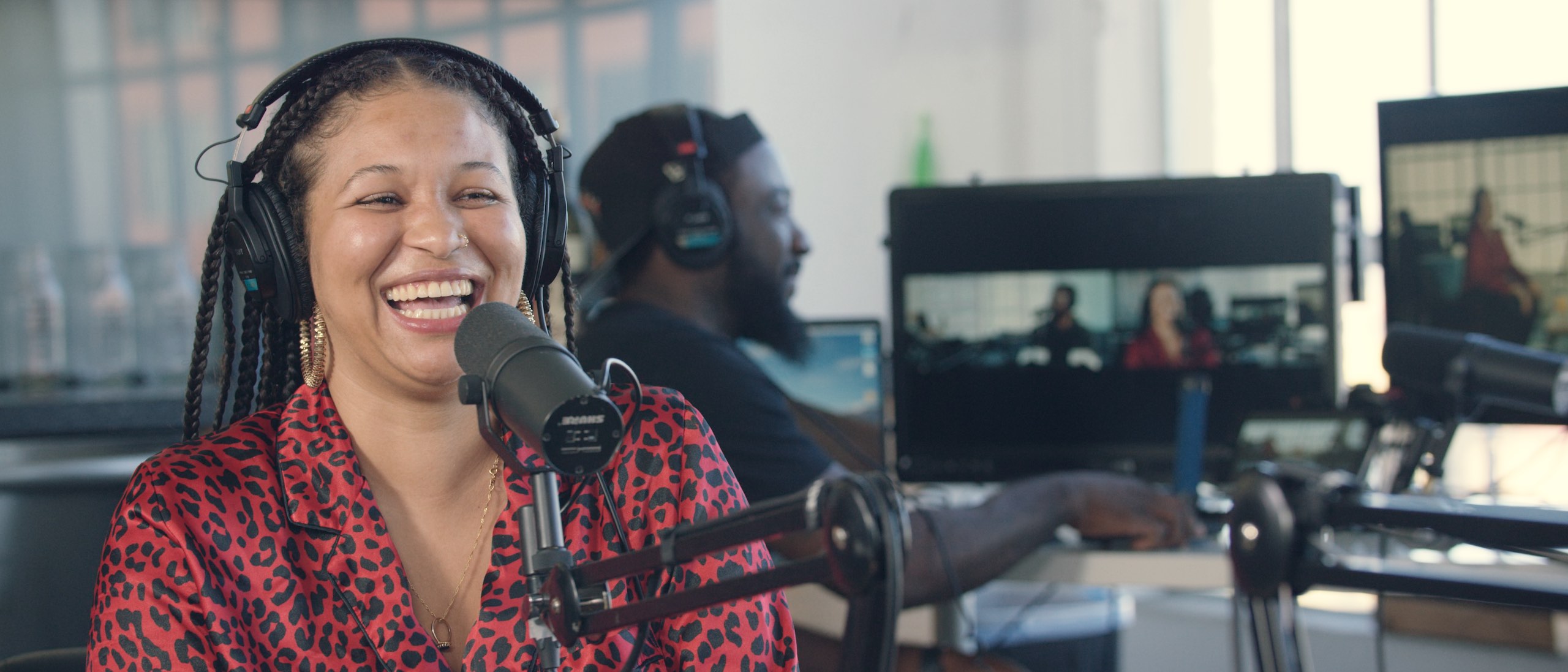 Uncreative Radio with Gabi Duran with braided black hair wearing black headphones laughing by microphone with side profile of African American man in the background by computer also wearing black headphones