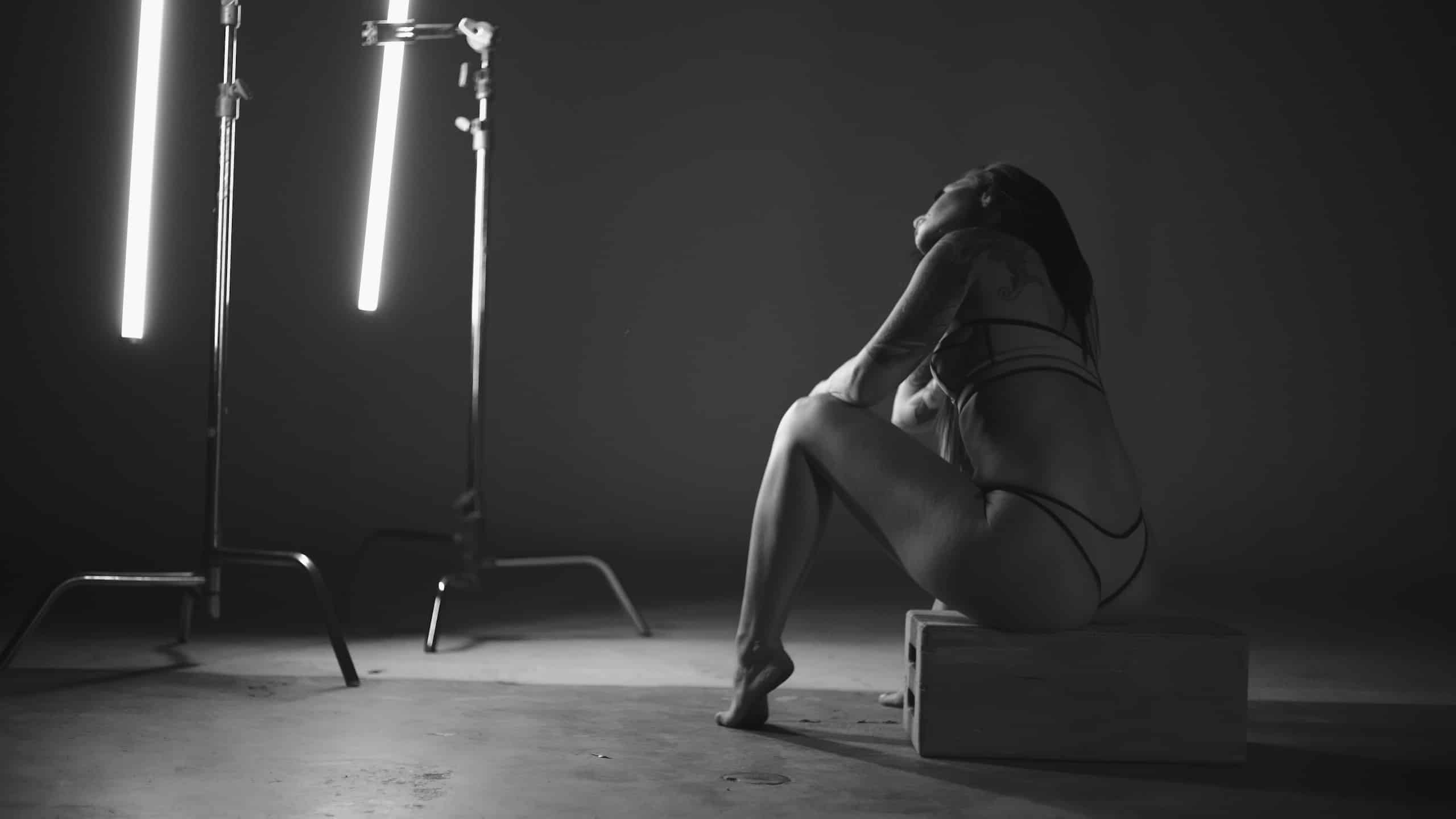Black and white view from behind of tattooed woman Jaquelyn Puma wearing bikini and thong sitting on a wooden box under lights