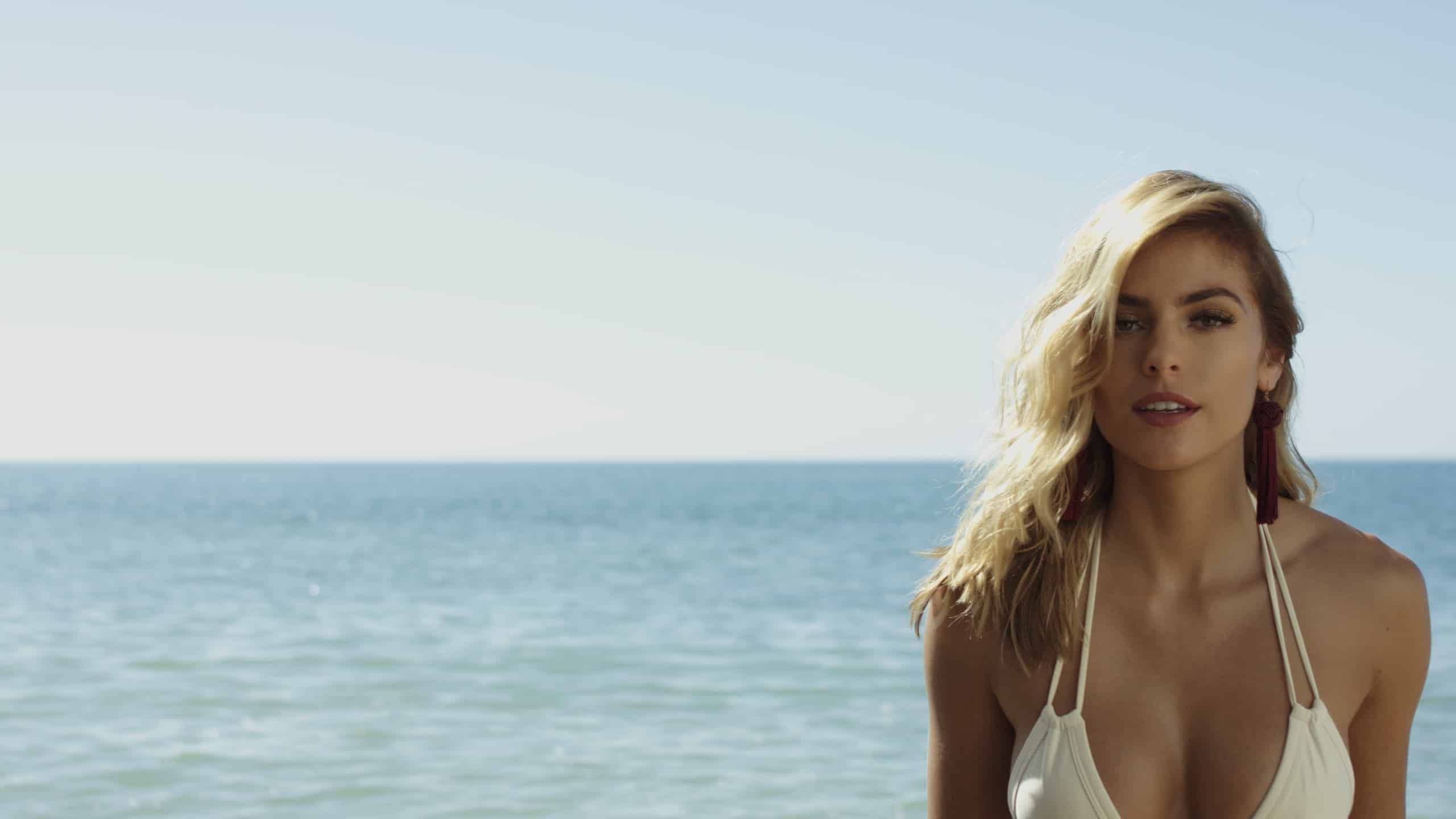 Dailies 0058 BTS with Montce Swim Woman with long blond hair wearing a white bathing suit posing for the camera by water