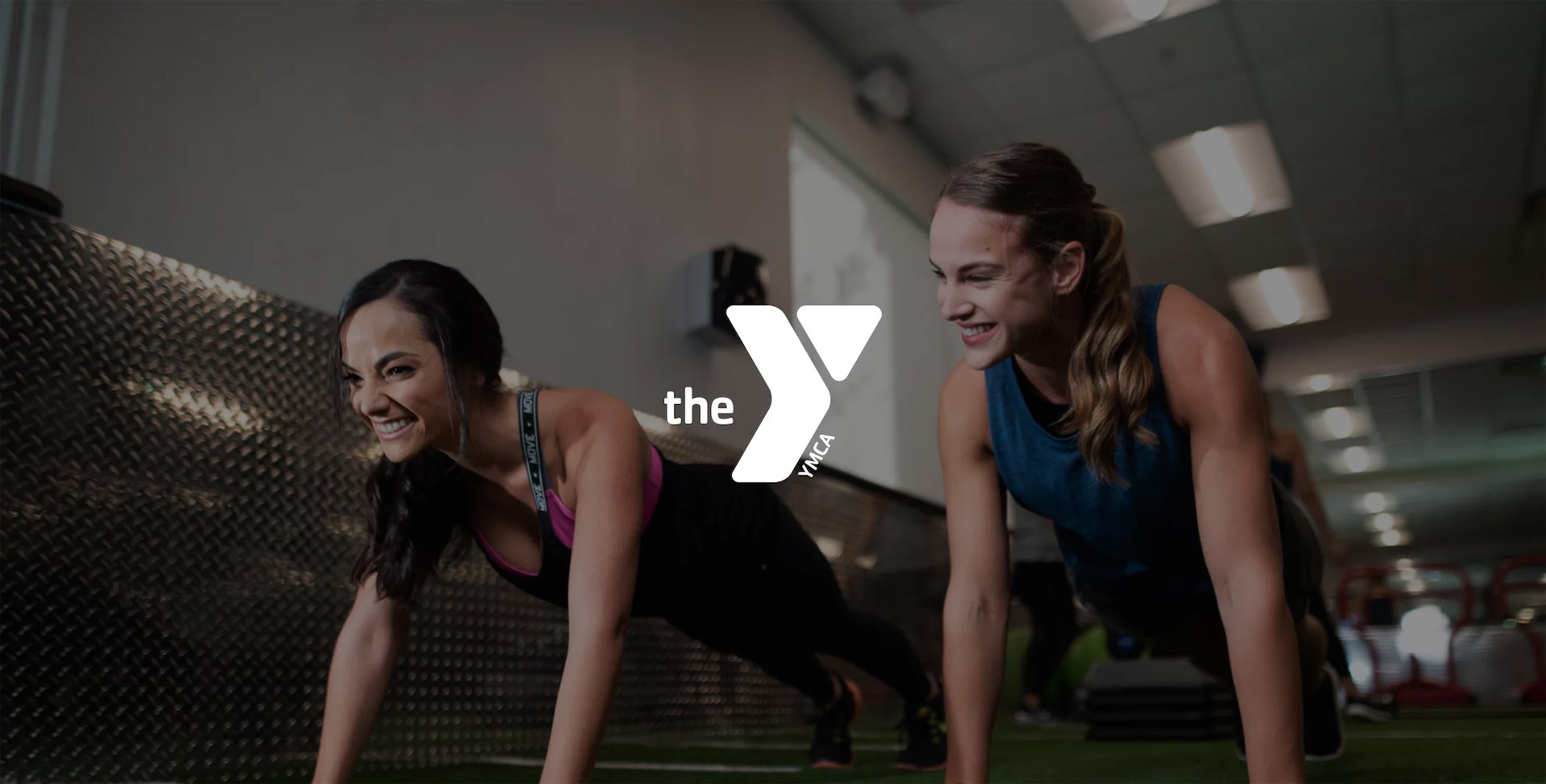 White The YMCA logo with two smiling women in plank poses in a gym