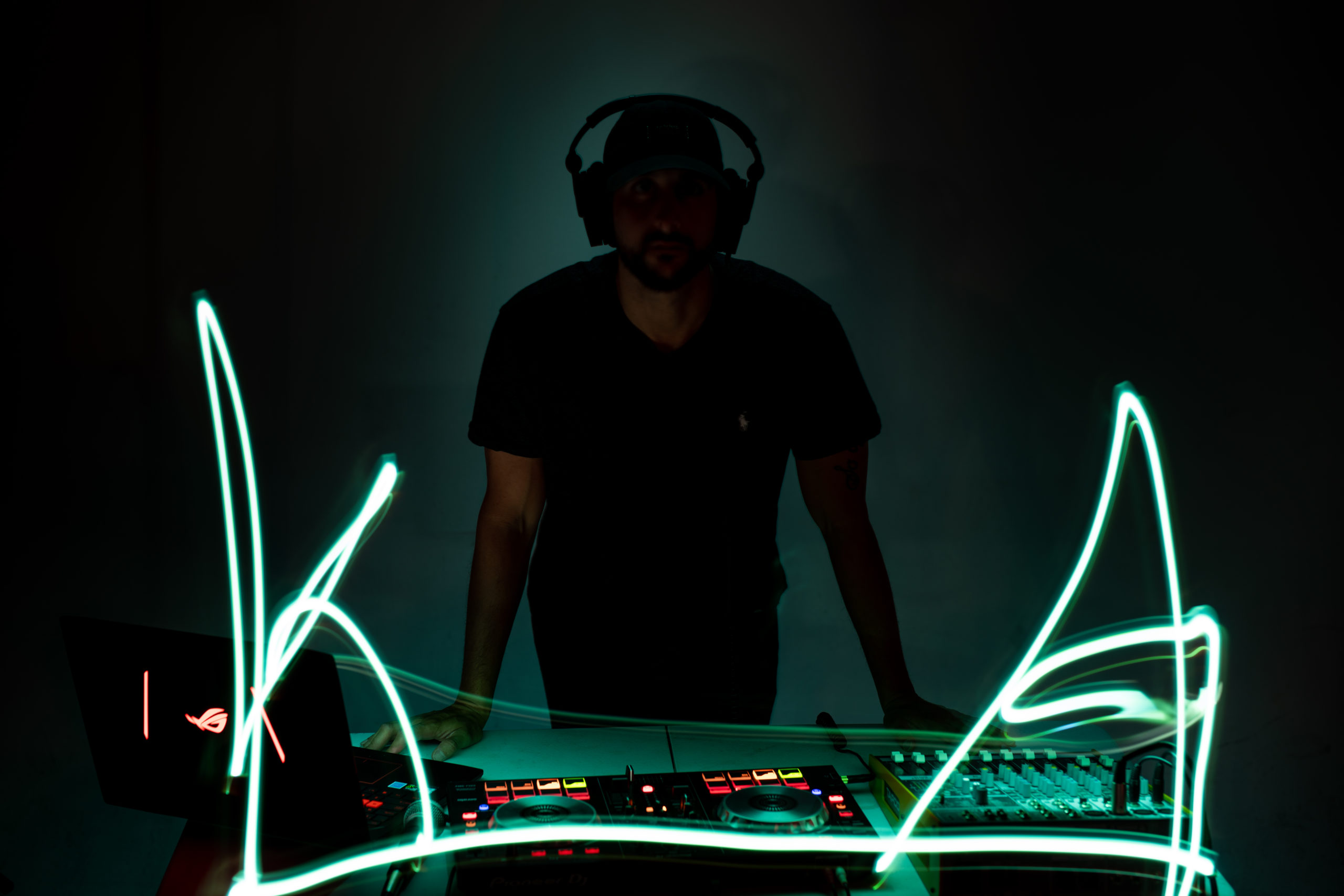 The importance of 3d animations-Male standing with black hat, beard and clothes looking towards camera behind a DJ machine with green neon lights. He is wearing headphones.