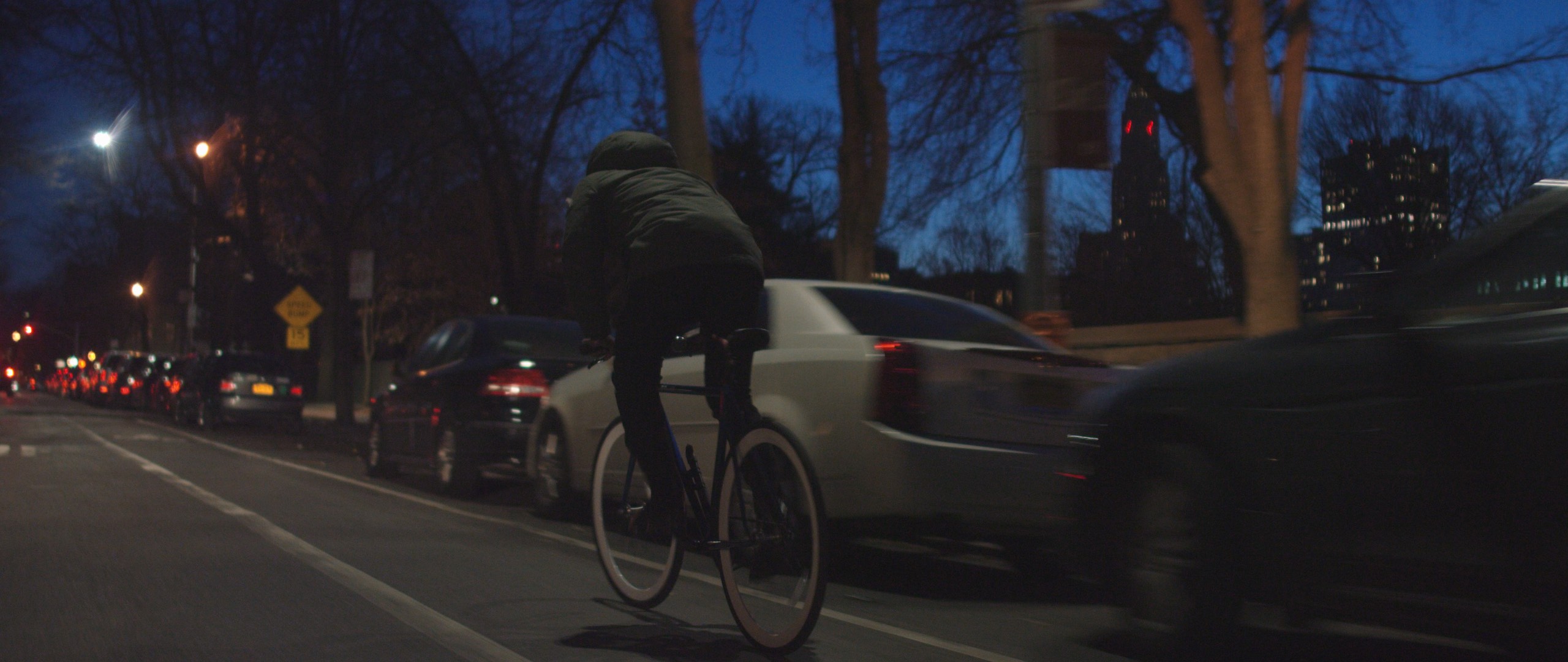 Christmas Eve Film C&I Studios View from behind of person on bicycle with white rimmed wheels biking down the street past cars at dusk
