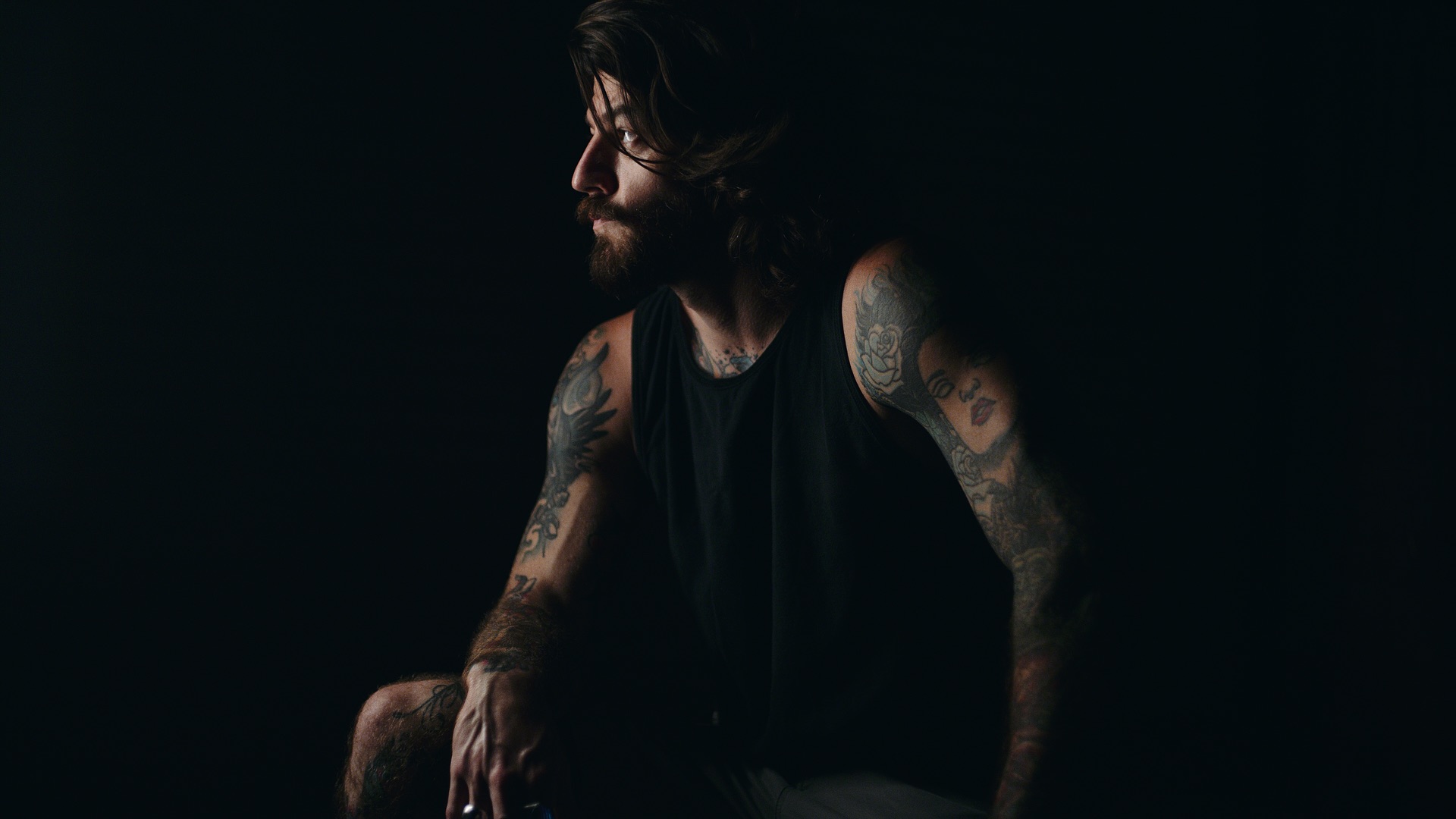 Luke Adams Crew Call the Tattoo Project Promo Man with beard and tattoos wearing a black tank top posing for the camera in semi darkness looking off to the side