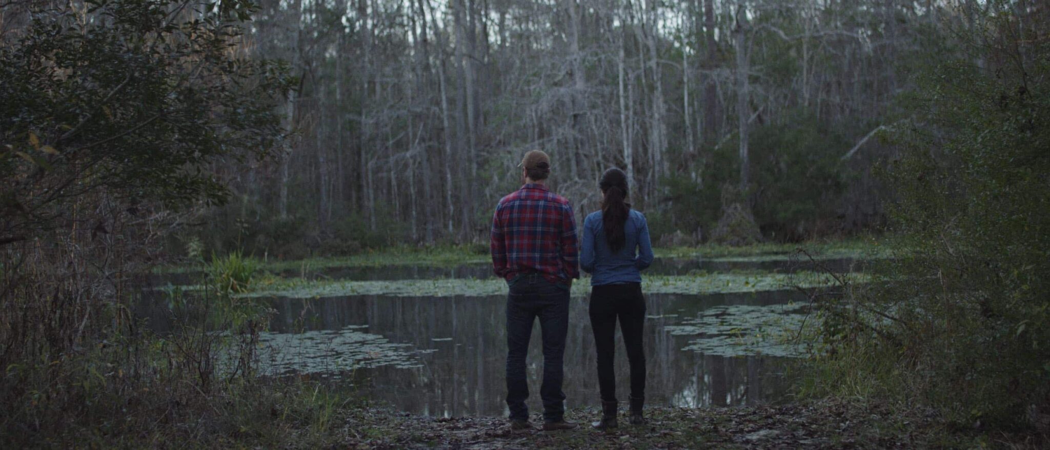 Dailies Season 1 Episode 6 The River Cabin Film View from behind of man wearing brown cap and flannel shirt with jeans and woman wearing a blue sweater and jeans standing by a lake