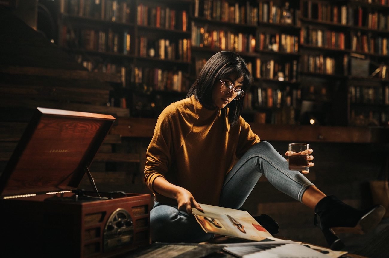 IU NARCITY Media This Hidden Cozy Coffee Shop In Fort Lauderdale Is A Bookworm’s Haven Woman with short black hair wearing glasses sitting on the floor next to an old vinyl player holding a glass of alcohol looking at brochure