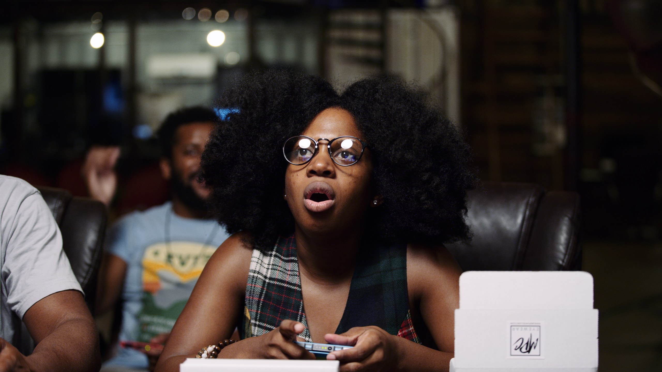 Reach Out and Play Woman with fizzy hair wearing glasses using a video game controller and responding to a video game that is being played
