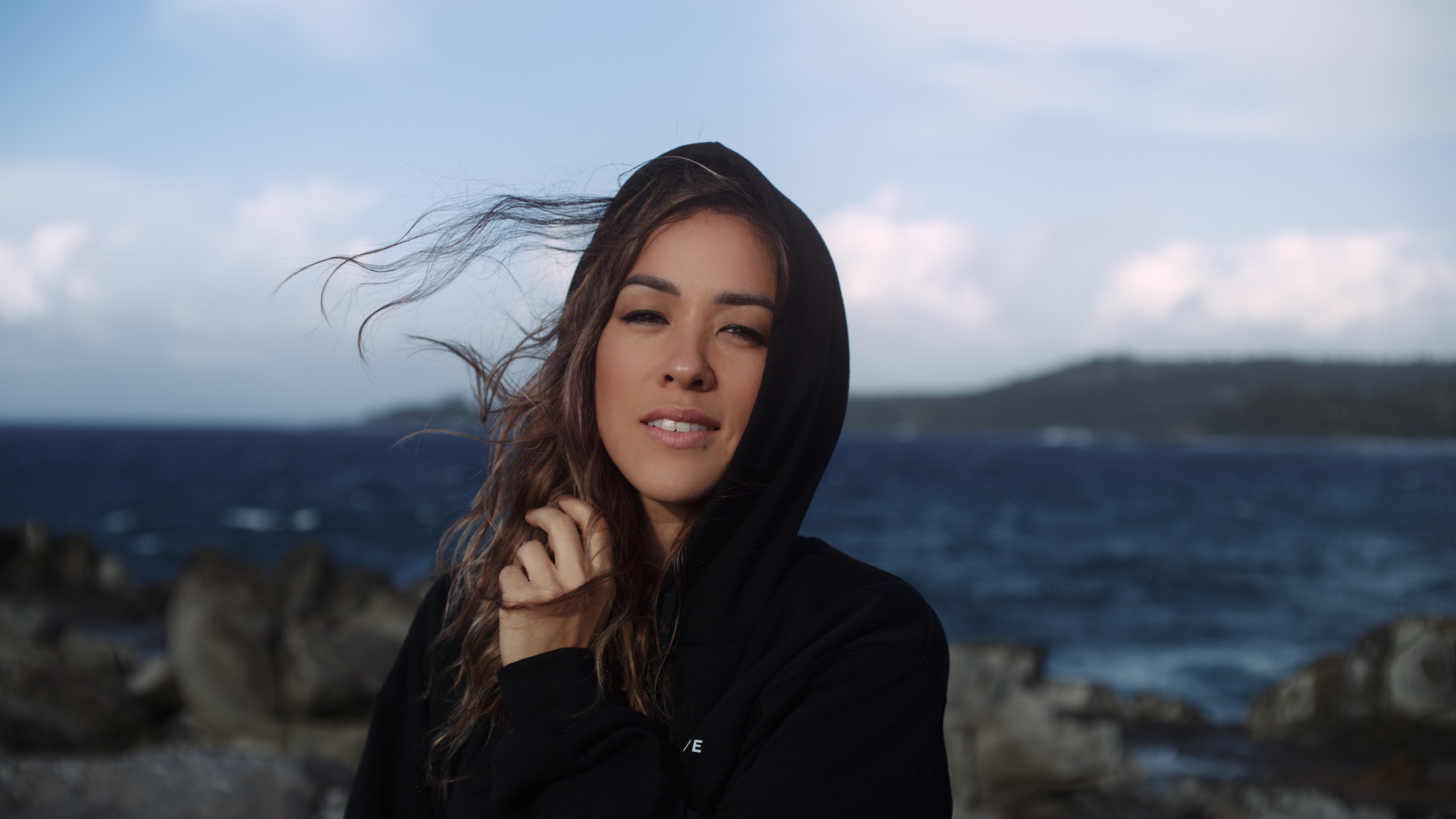 Uncreative Shop Hawaii Apparel on available at the Uncreative Shop Headshot of woman with long brown hair wearing black hoodie posing for the camera by a lake