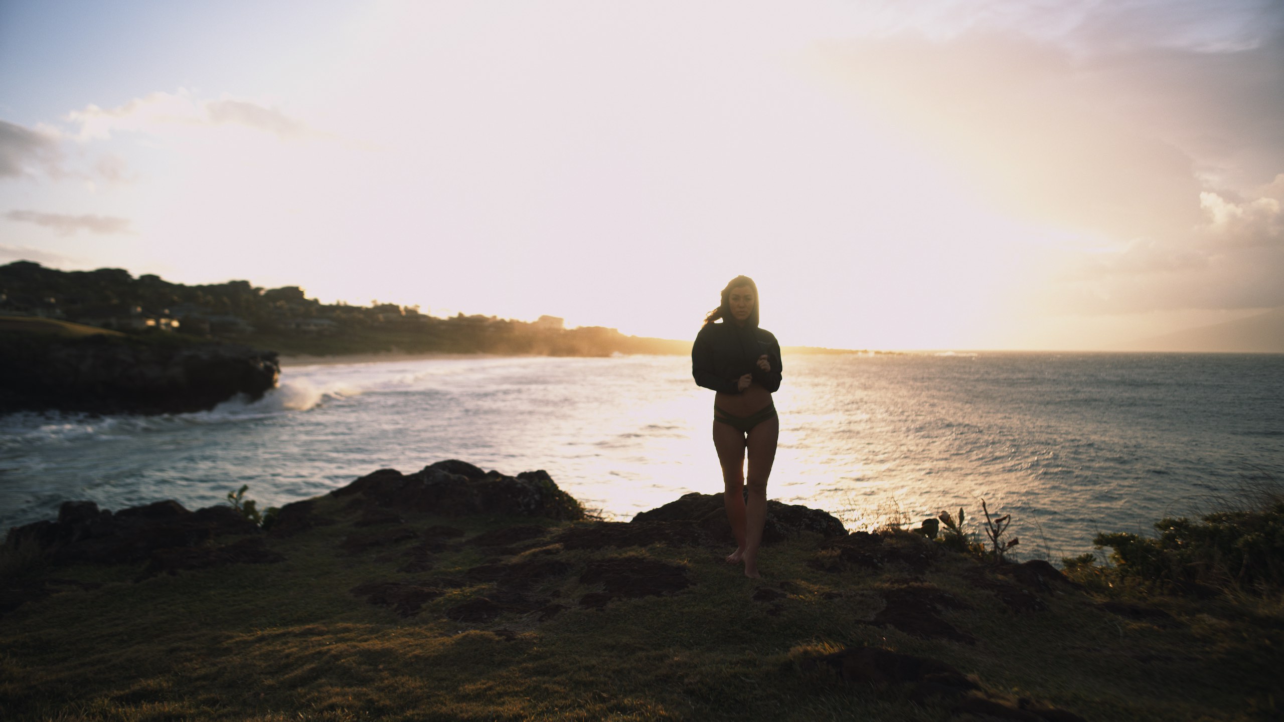 Uncreative Shop Hawaii Woman with long hair posing with a hoodie and bikini bottom by water with the sun setting behind her