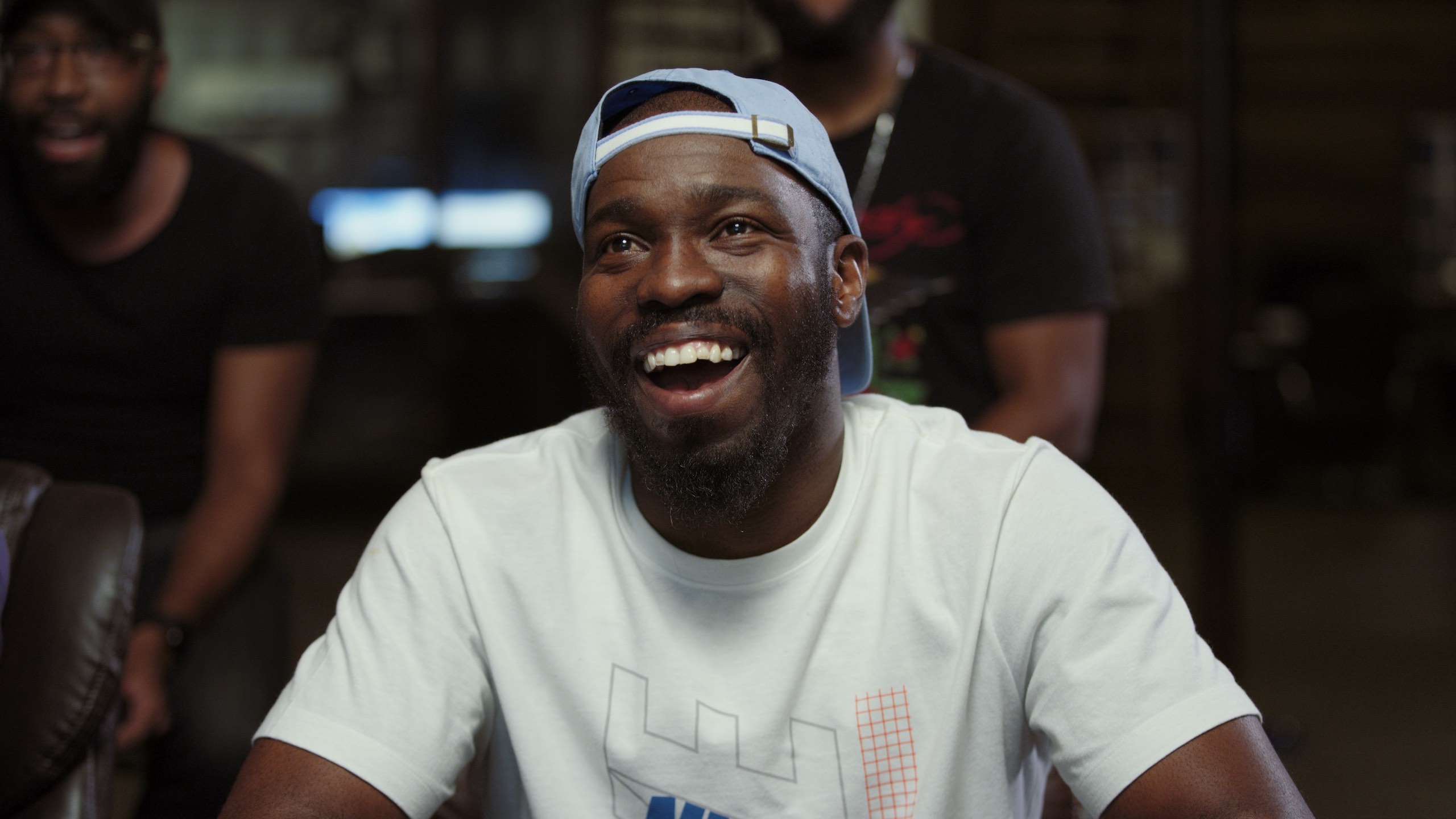 Three men playing and watching a video game in session with one bearded African American man wearing light blue cap backwards and white t shirt