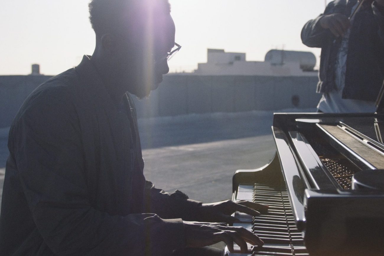 Side profile of man playing a piano on a rooftop with the sun shining on him with man nearby playing a violin