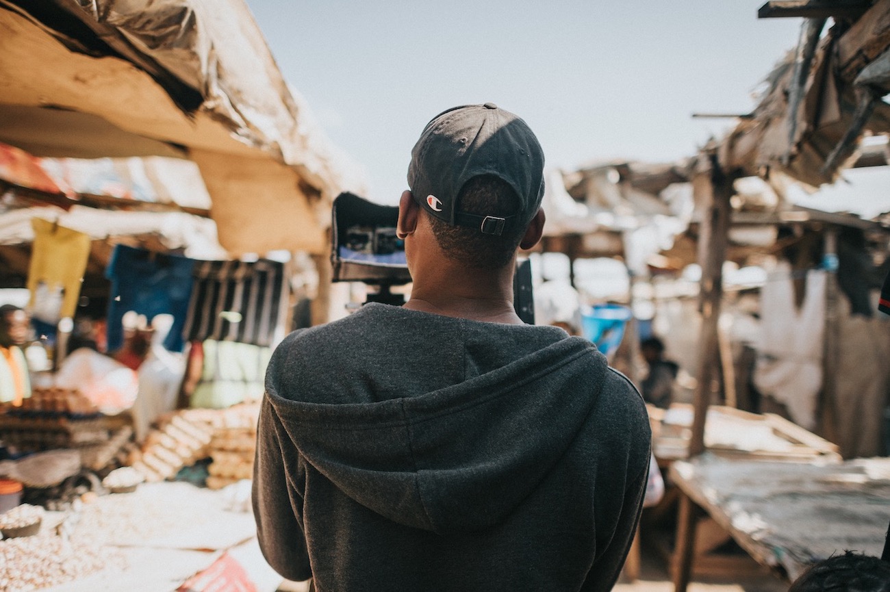 View from behind of him wearing a gray cap and sweatshirt filming a scene in a market