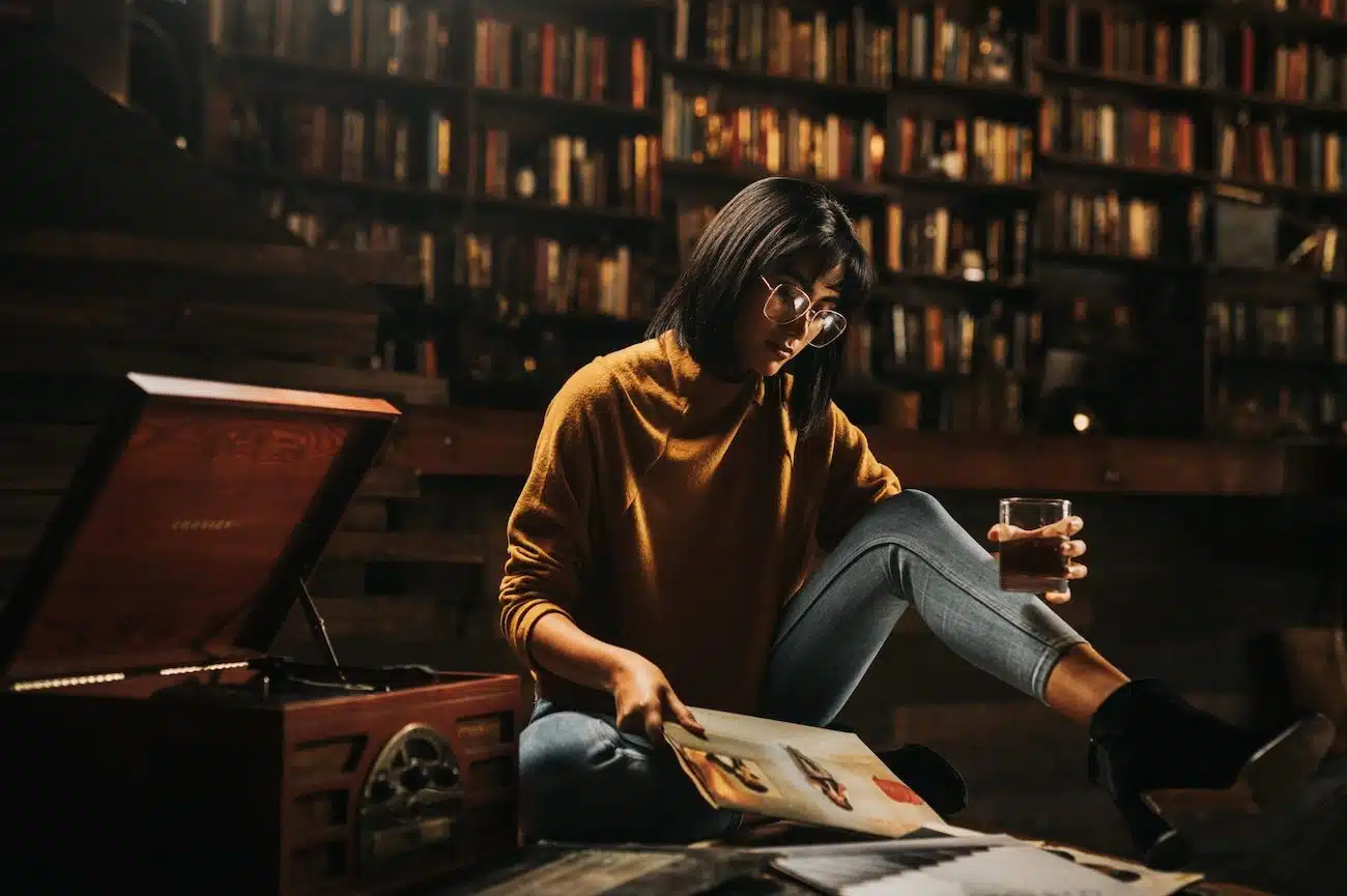 Woman with short black hair wearing glasses sitting on the floor next to an old vinyl player holding a glass of alcohol looking at brochure