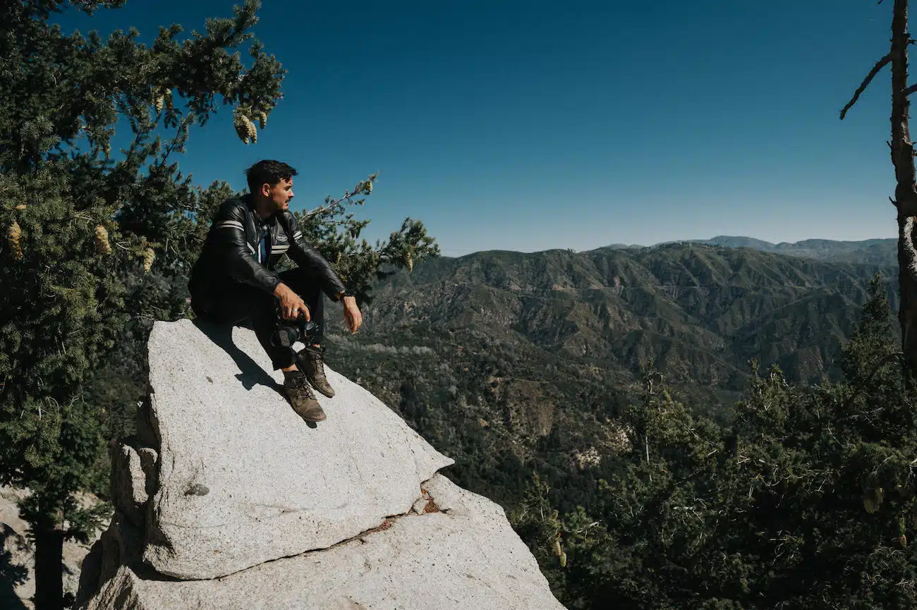 Man wearing black leather jacket and brown leather boots sitting on a white rock looking over the valley