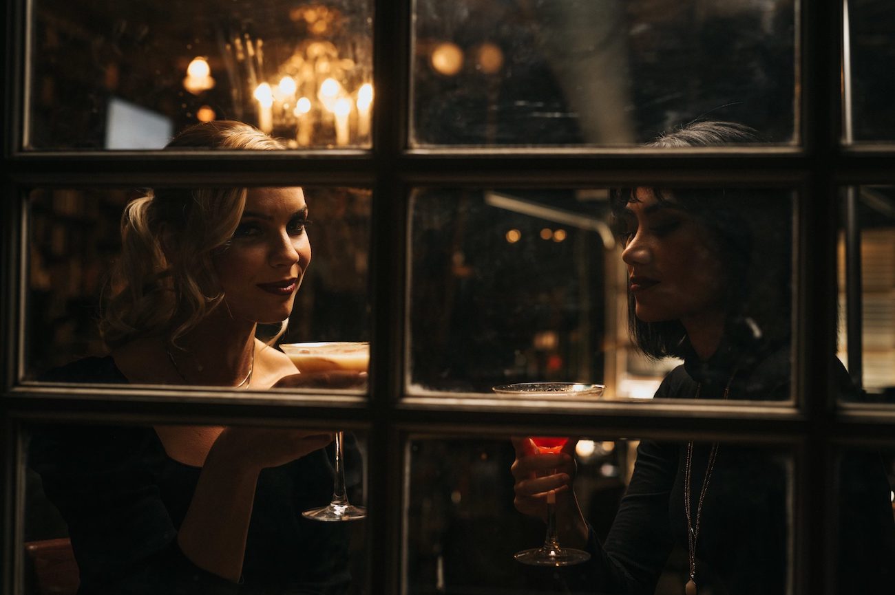 Two woman holding martini glasses looking out a window