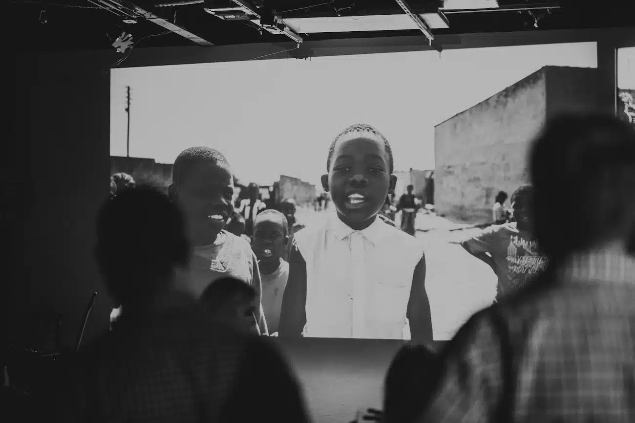 Black and white view from behind of group of people looking at screen with a movie playing on it