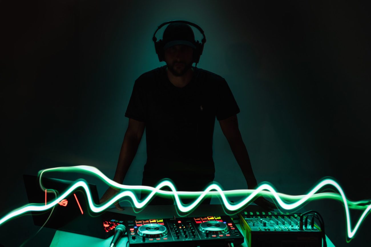 Silhouette of man wearing cap and headphones standing behind DJ equipment lit up by green neon light