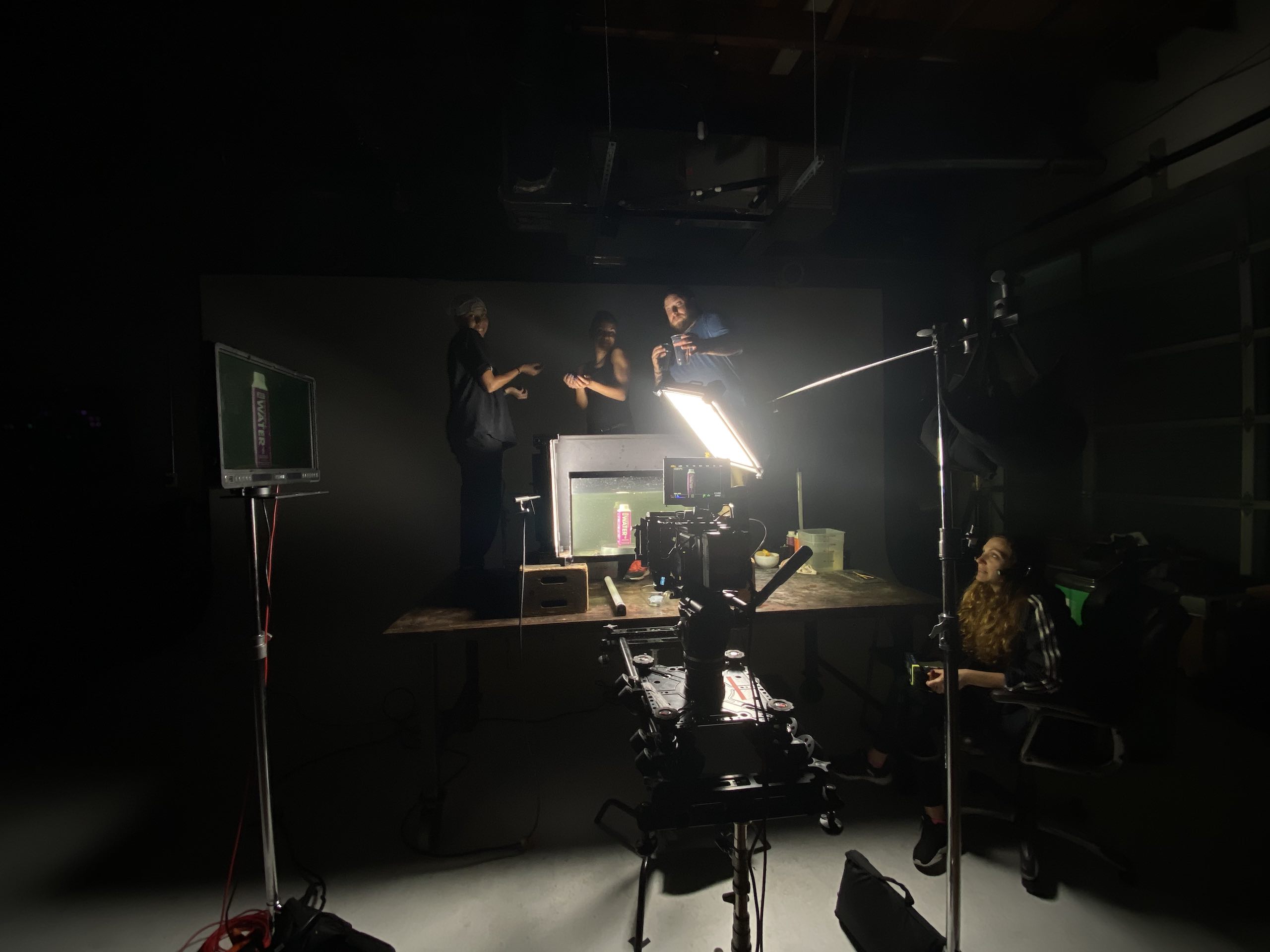 View from behind of camera trained on purple drink container in a tank filled with water in a studio with crew members standing and sitting by surrounded by lighting equipment