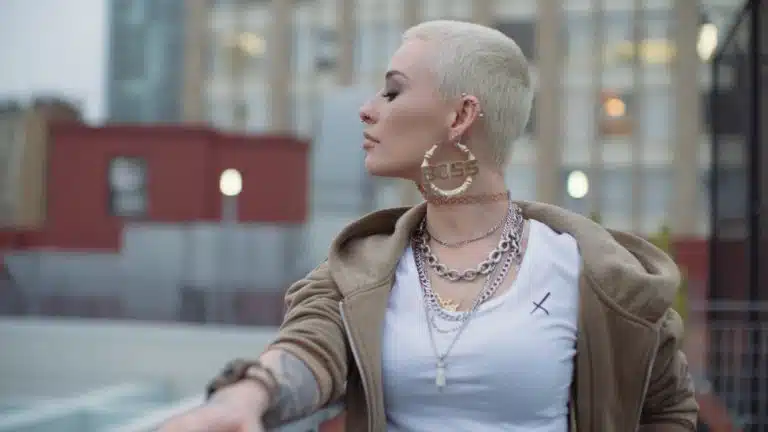 Woman with short blond hair wearing jewelry as well as white dress shirt and beige hoodie looking off to the side over her shoulder