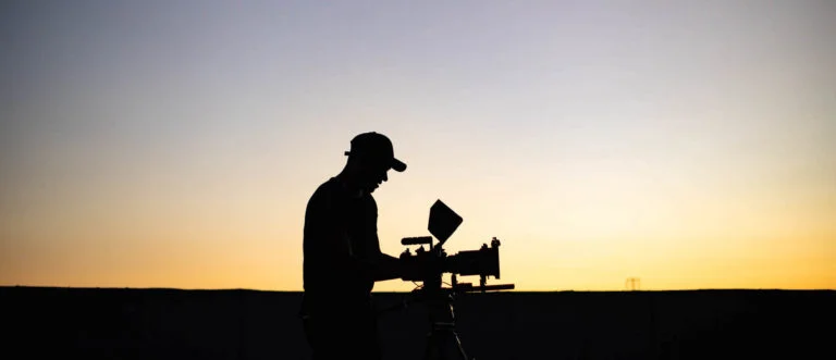 Keyword: Video Production Gear List .Silhouette side profile of crew member wearing cap using video camera at dusk