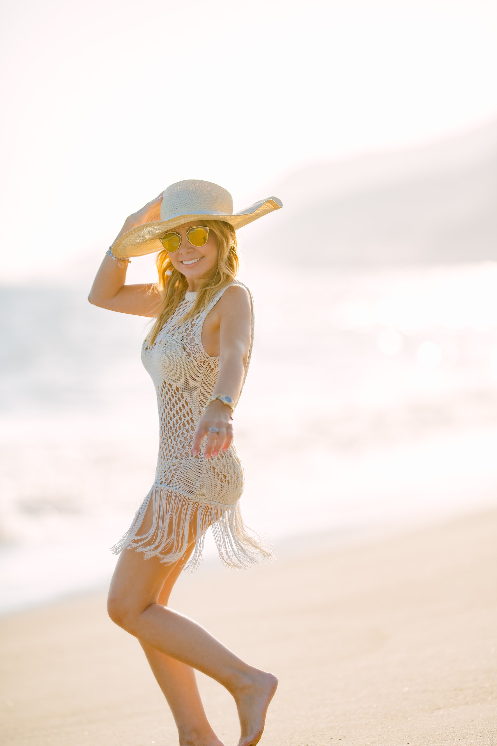 Peretti Italy Woman with long blond hair wearing a knitted white dress and a beige hat smiling for the camera on a beach barefoot