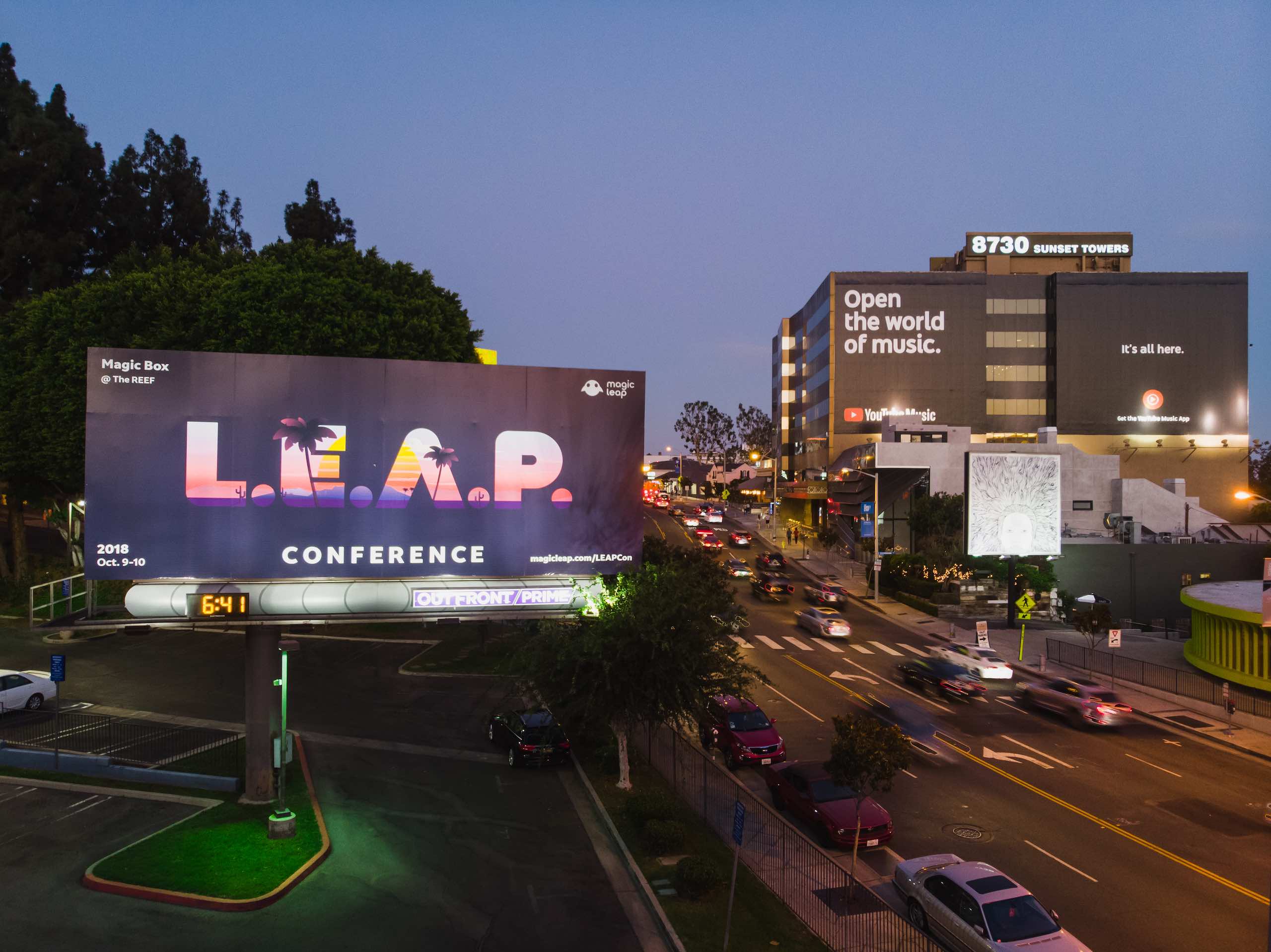 LEAP Conference billboard on display by a parking lot in city near road with cars parked and driving by at night