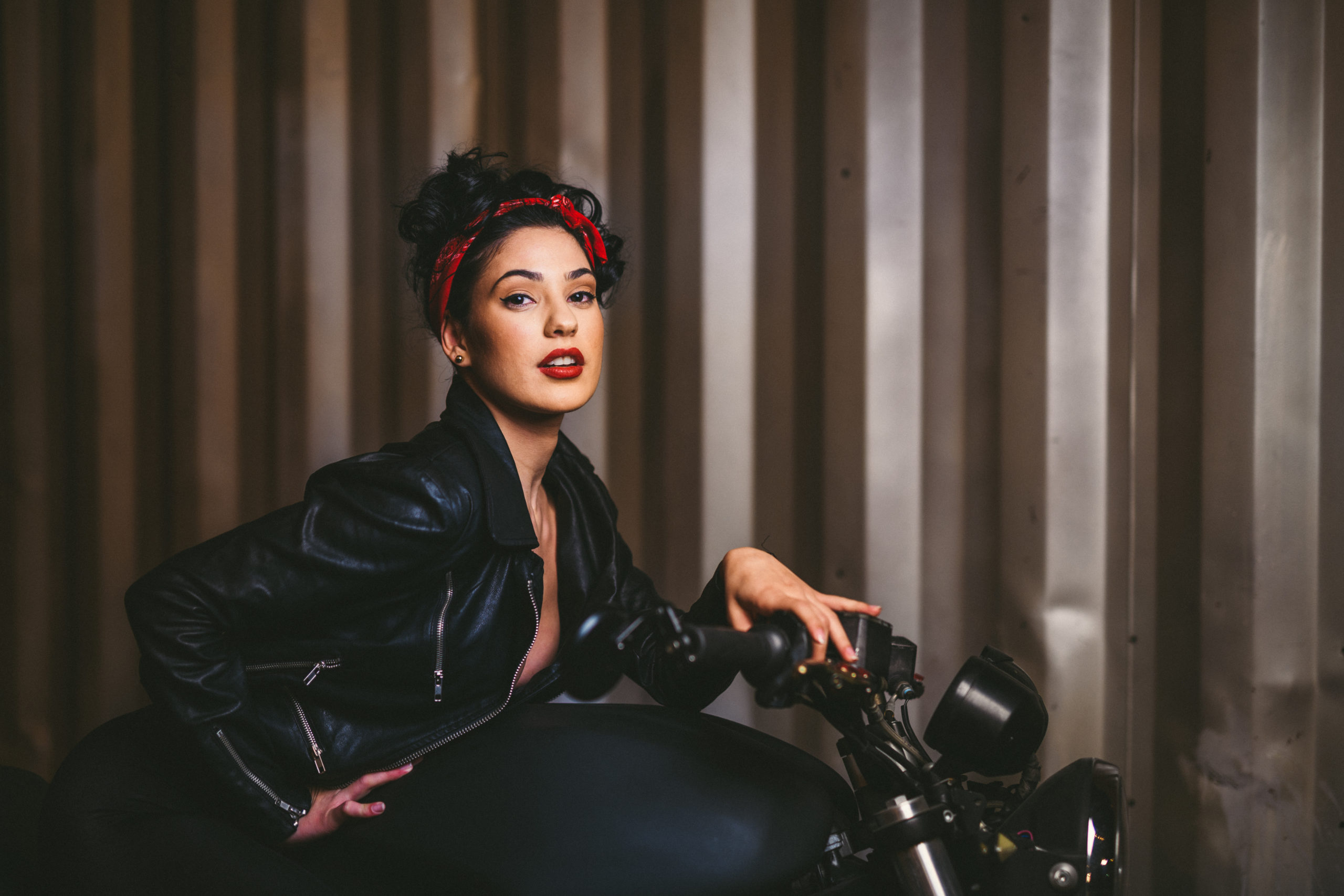 Closeup of woman with short black hair and red lipstick wearing a black leather jacket and red bandana posing for the camera sitting on a motorcycle