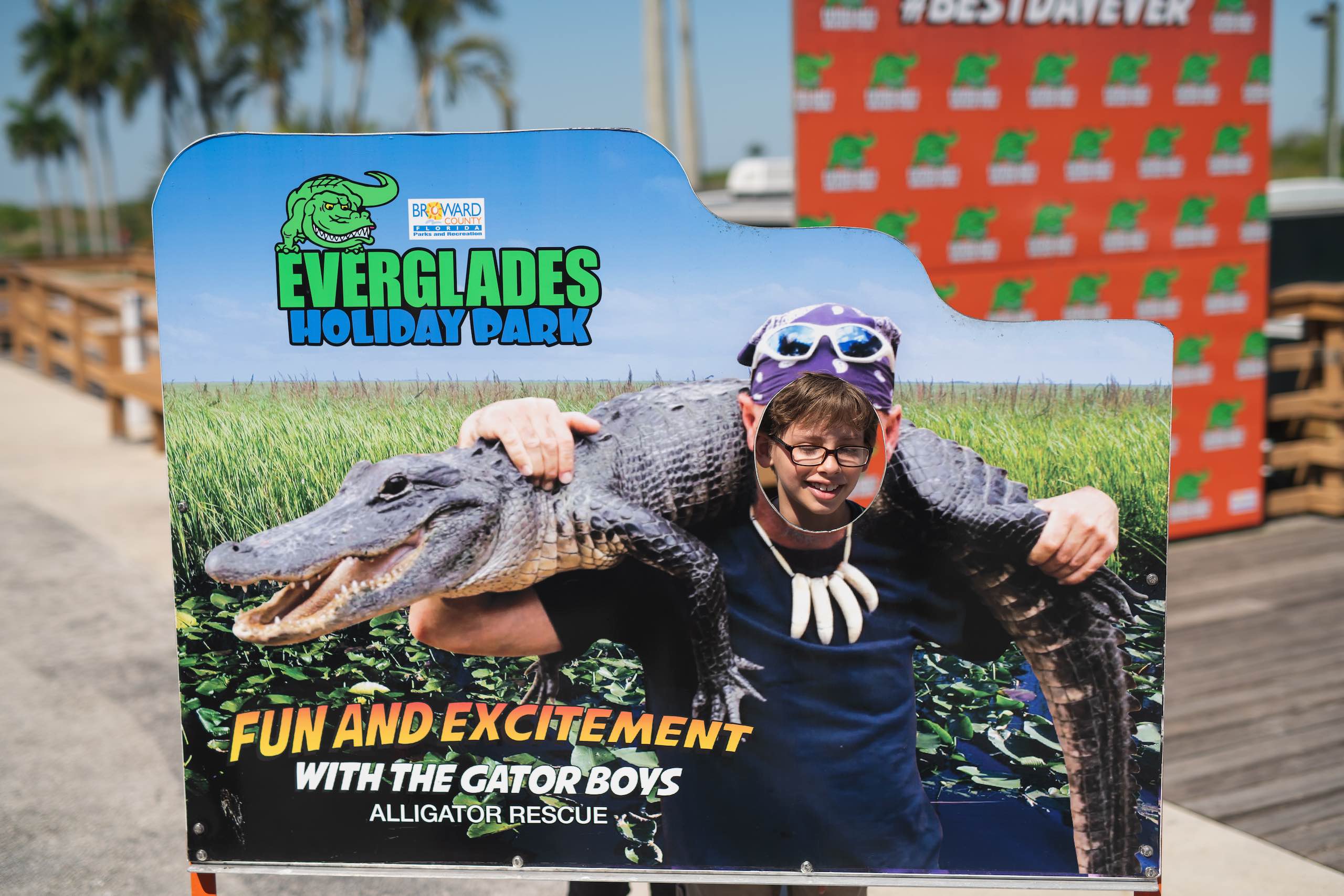 Everglades Holiday Park Closeup of boy looking through hole of a cutout of someone holding an alligator in his arms over his shoulders