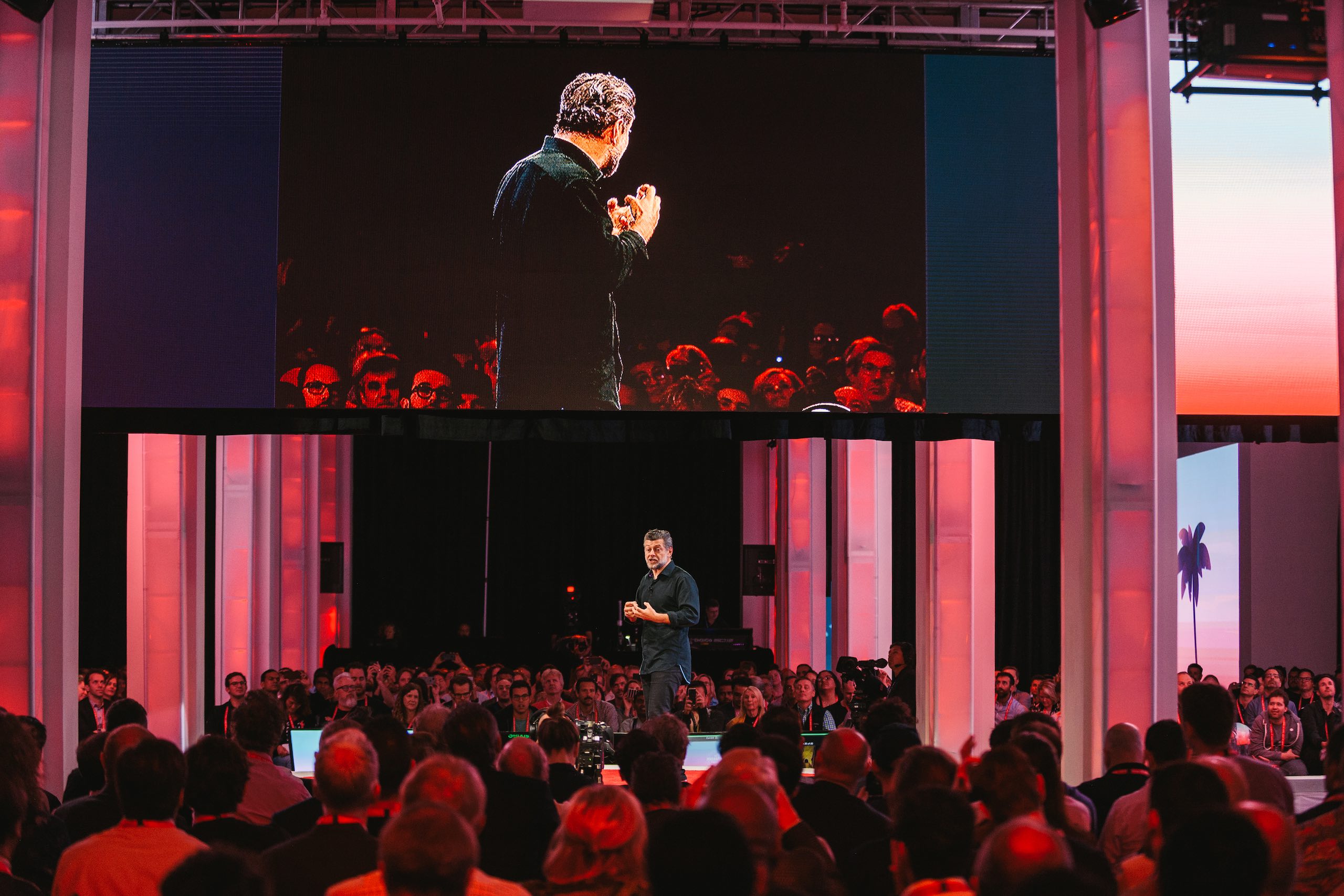 Man with short gray beard on stage at a Free Your Mind event talking to the audience surrounding him with him projected on a screen above him