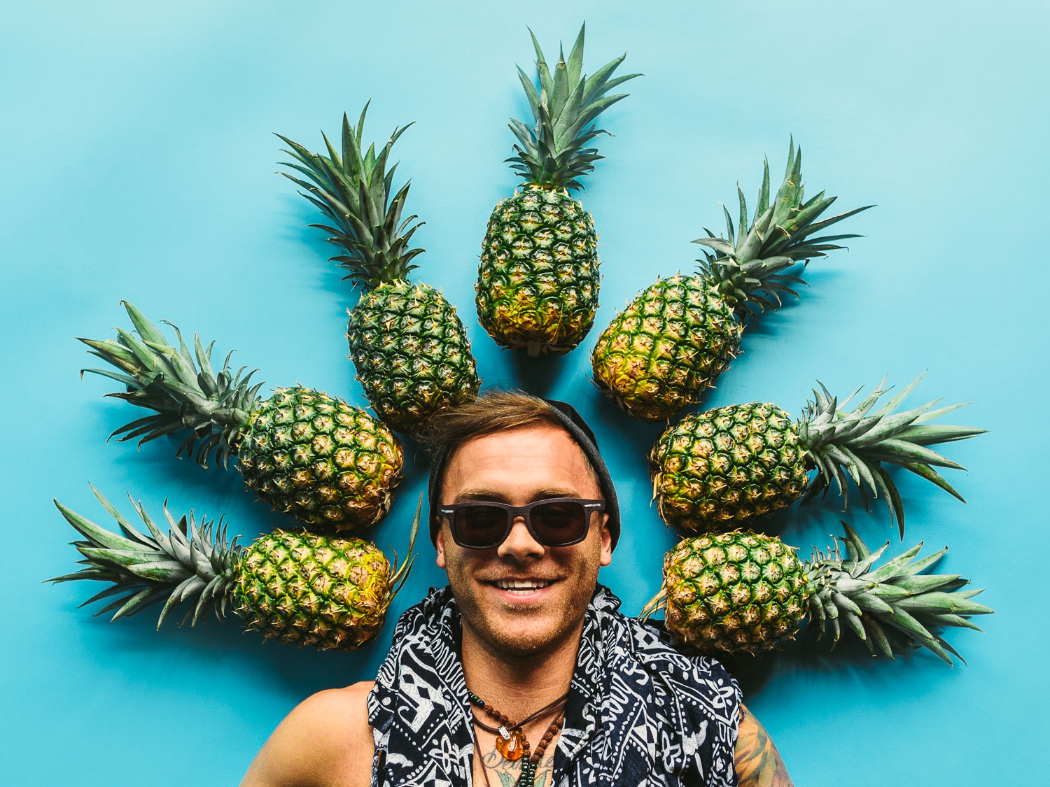 Artwalk 2018 May Pineapple Pyramid Man wearing a black knit cap smiling posing for camera with pineapples surrounding his head on a teal background