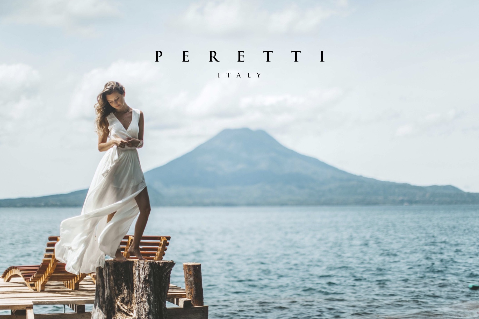 Peretti Italy Brand Guidelines Woman in white dress standing on a stump looking down by a pier and water with a mountain in the background