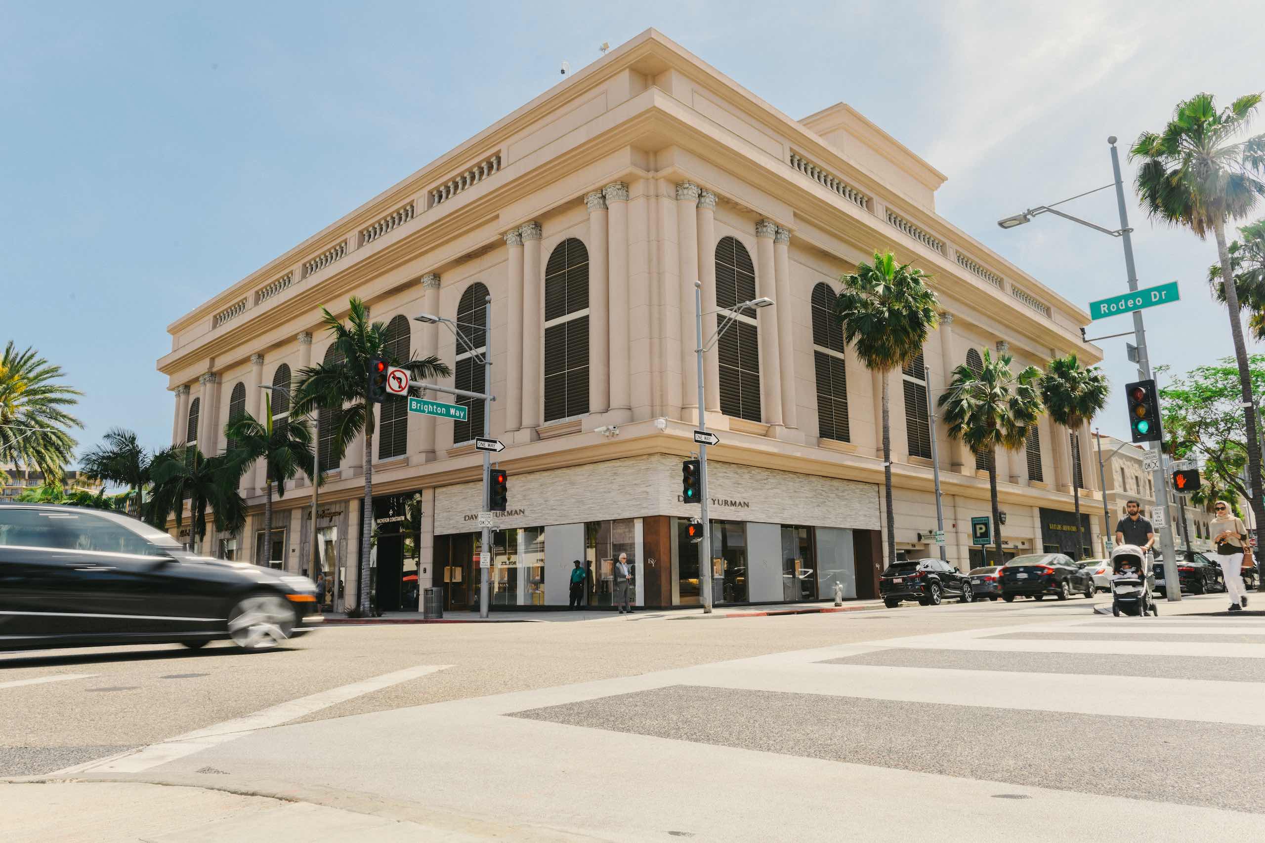 Peretti Italy Branding and Photography Building with palm trees at intersection of Rodeo Drive and Brighton Way with cars driving along the way and a man with a scroller in the crosswalk