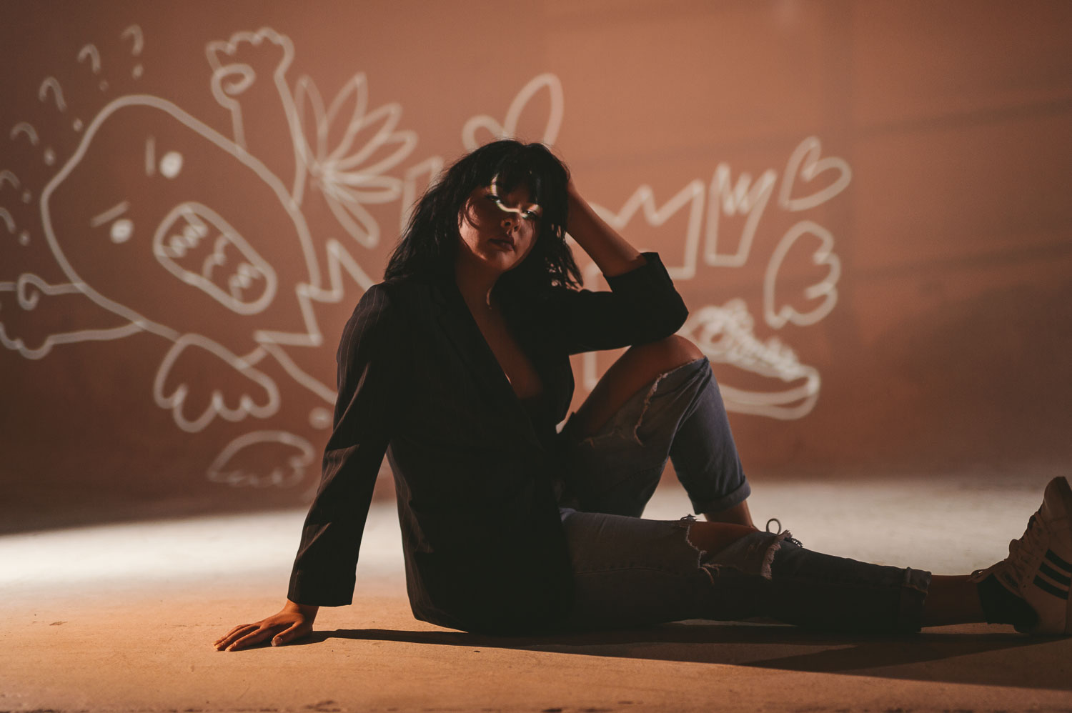 Artwalk Self Projection with woman with black hair wearing torn jeans and sneakers posing for camera sitting on the floor with part of white artwork being projected on her