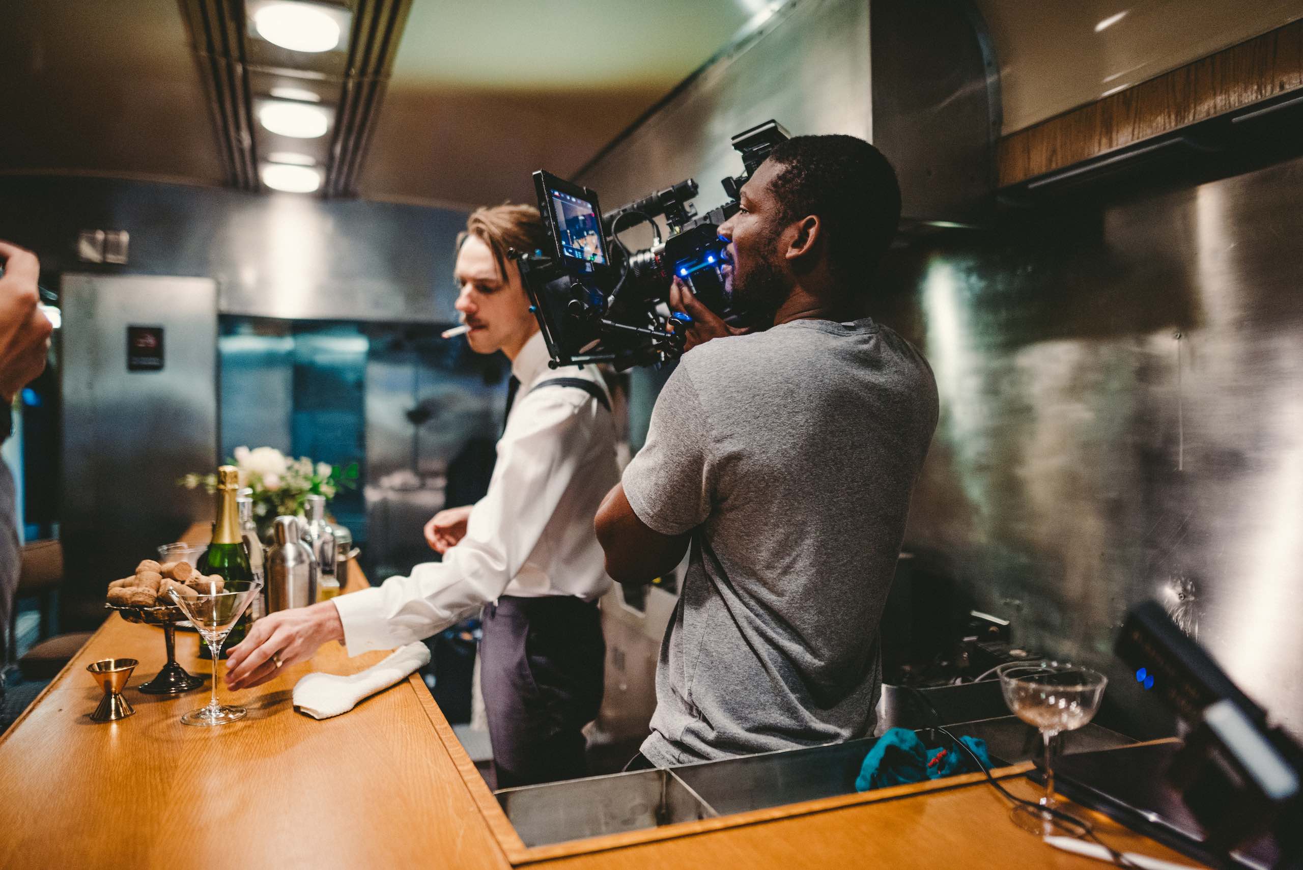 View from behind of Joshua filming a bartender with a cigarette in his mouth in a train car